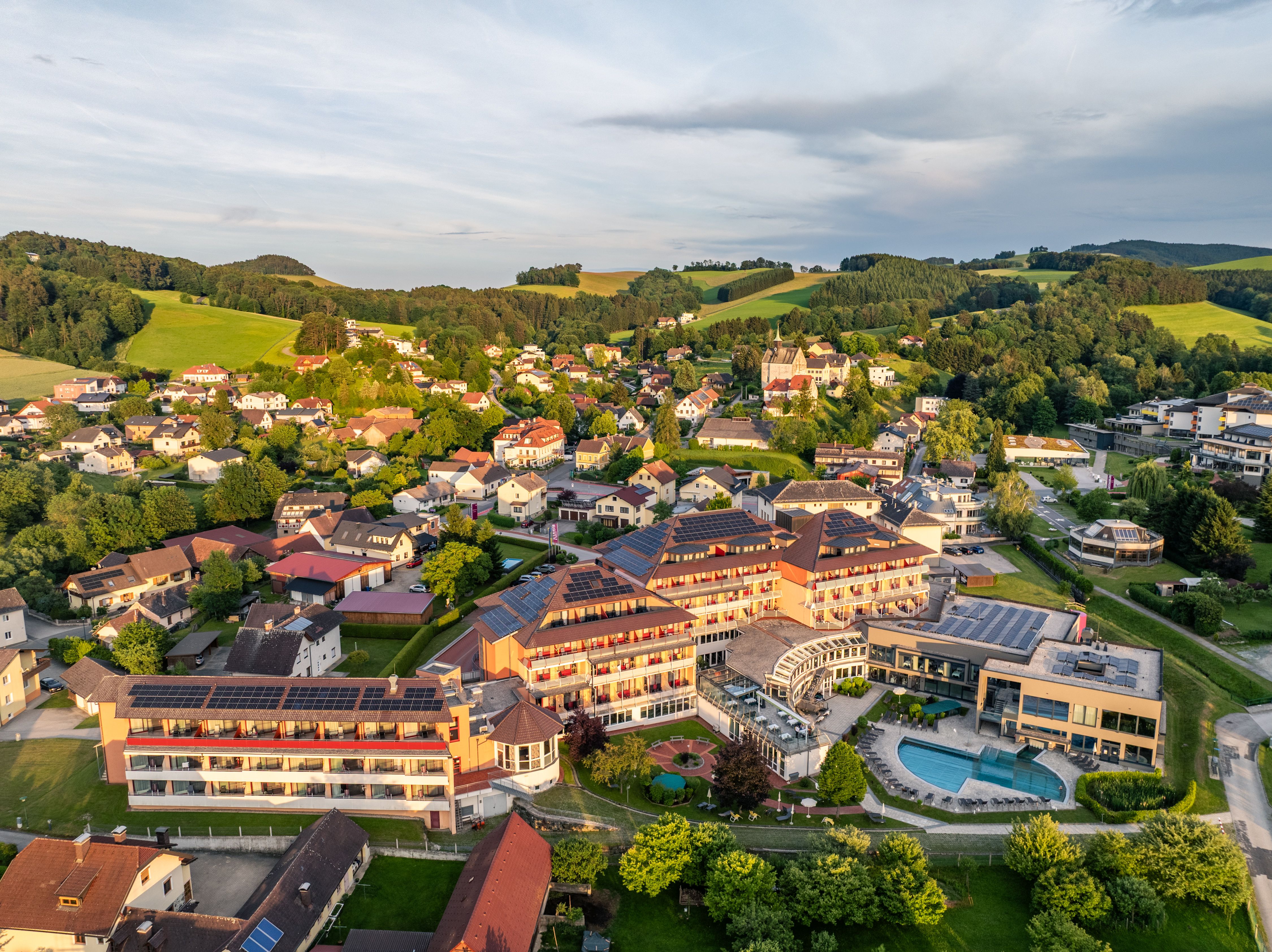 Letecký pohľad na Bad Schönau s veľkým hotelom s bazénom v popredí a zelenými kopcami v pozadí.