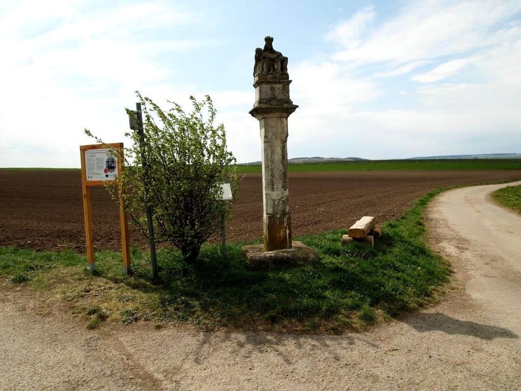 Kamenný stĺp pri poľnej ceste s informačnou tabuľou a kríkmi.