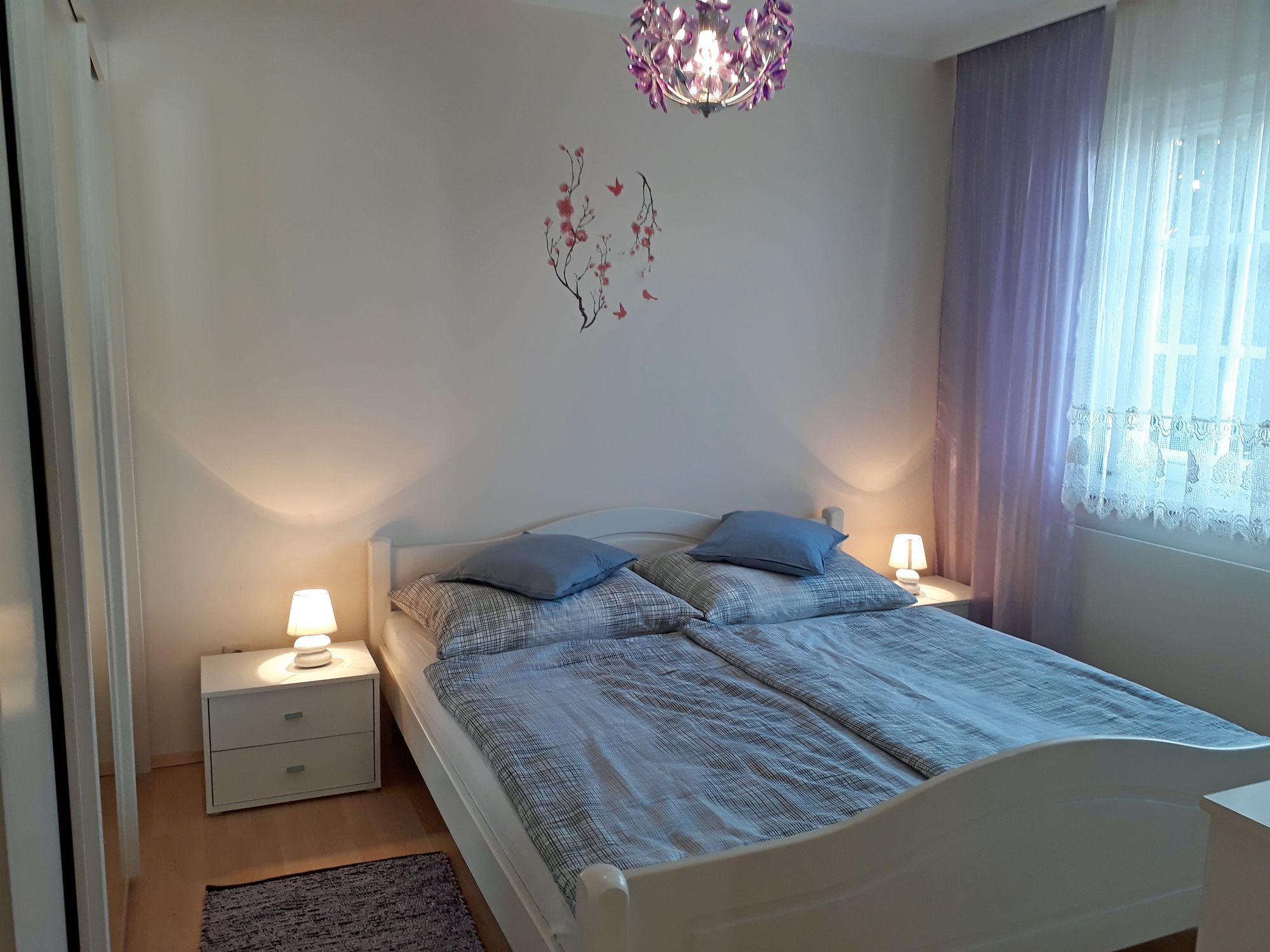 Útulná spálňa s manželskou posteľou, dvoma nočnými stolíkmi s lampami a fialovými závesmi.