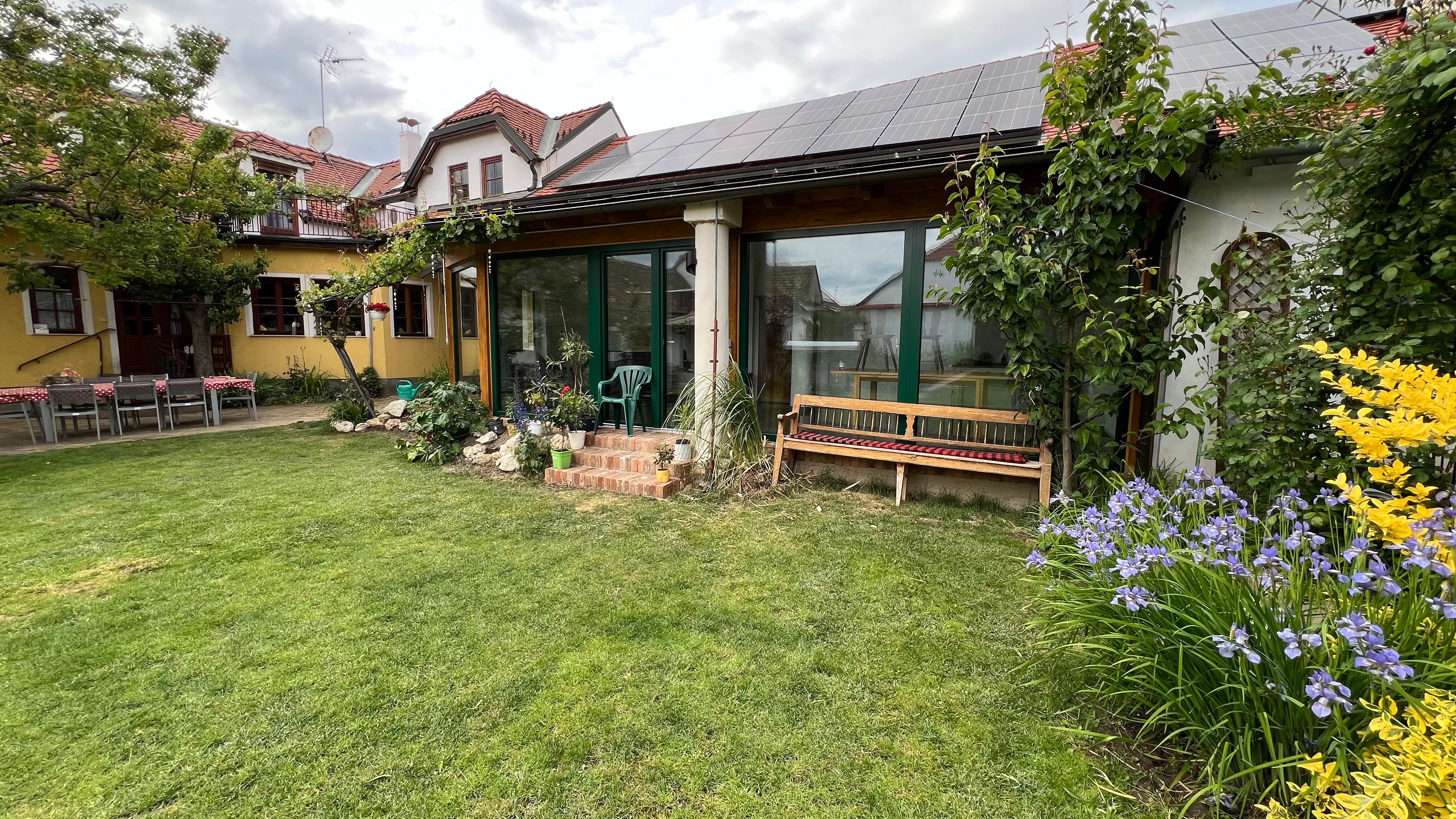 Záhrada s trávnikom, kvetinovými záhonmi a domčekom so solárnymi panelmi na streche.