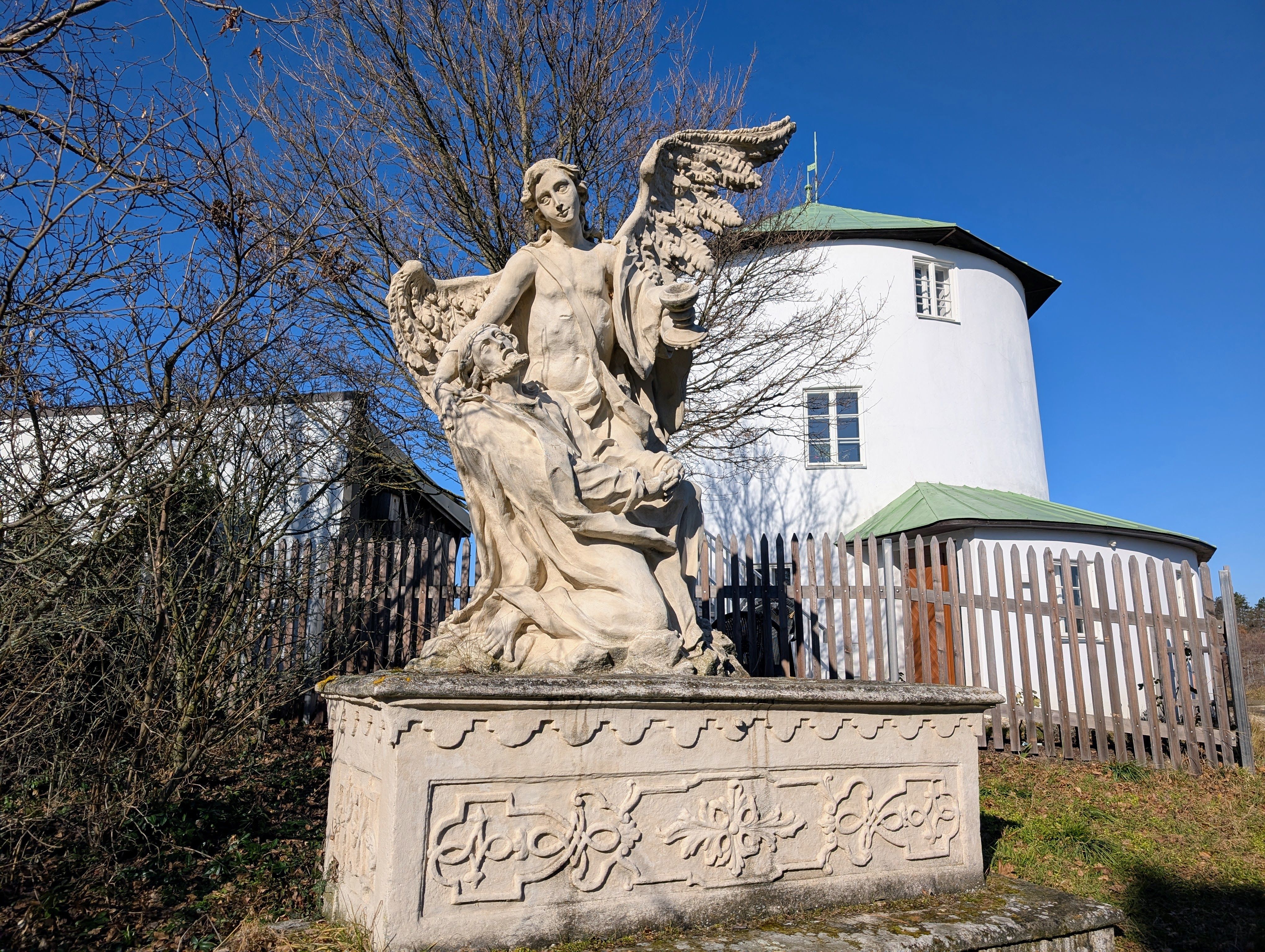 Kamenná socha anjela s krídlami a kľačiacou postavou pred okrúhlou bielou budovou so zelenou strechou.