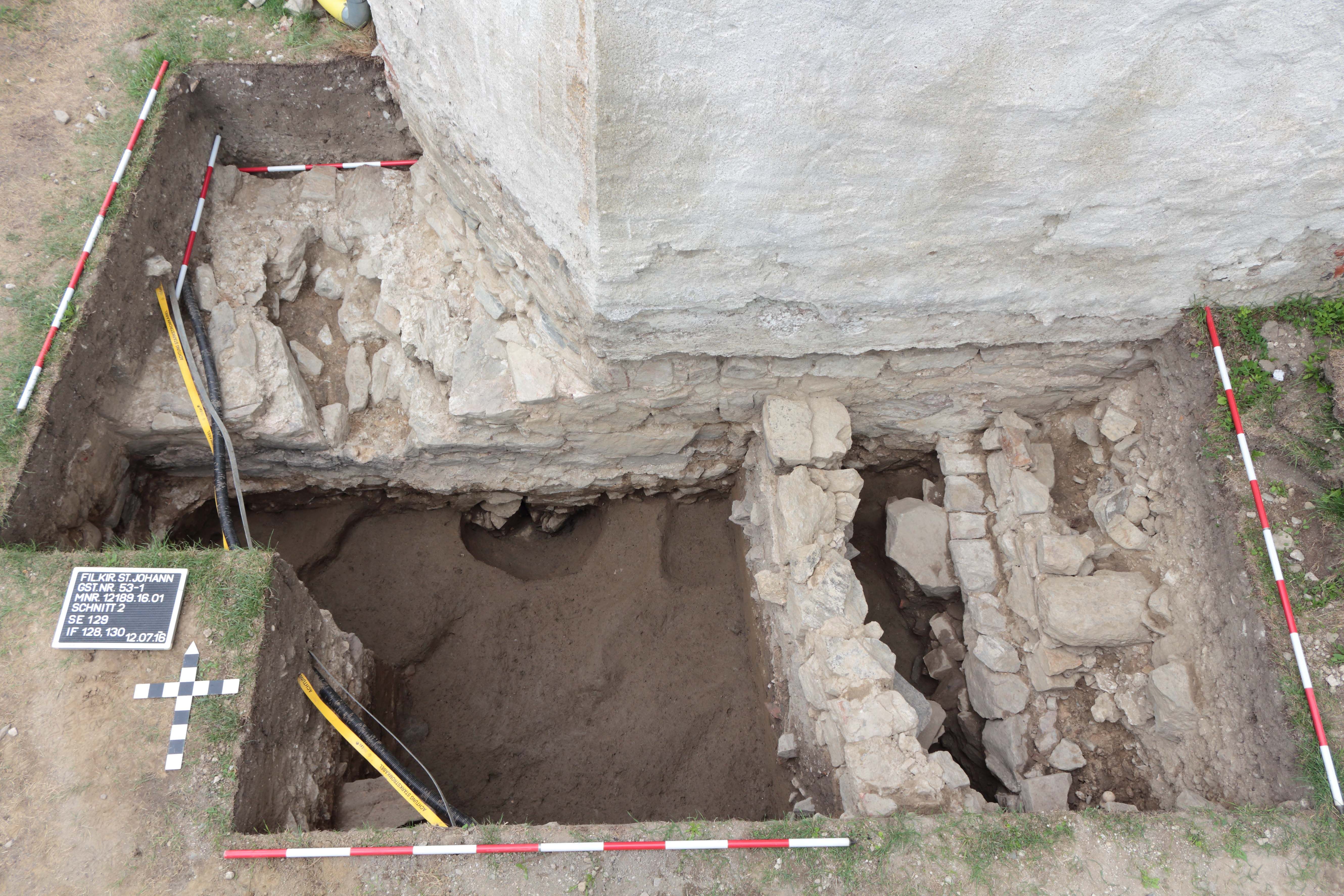 Archeologický výkop s odkrytými zvyškami múru a meracími tyčami.