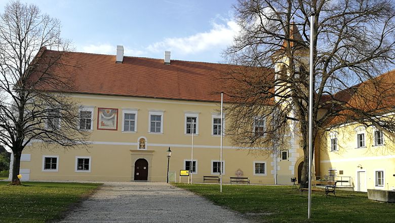 Žltá budova hradu s červenou strechou a vežou, obklopená stromami.