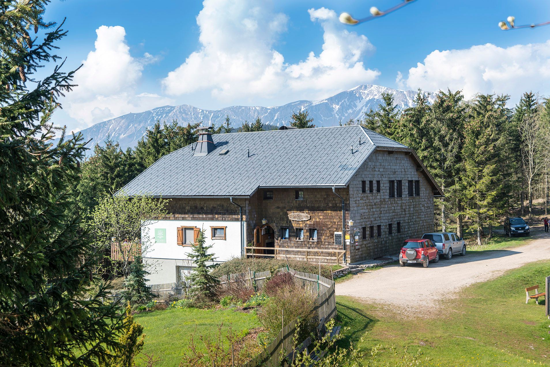 Rustikálny prístrešok v horskej krajine so stromami a autami v popredí.
