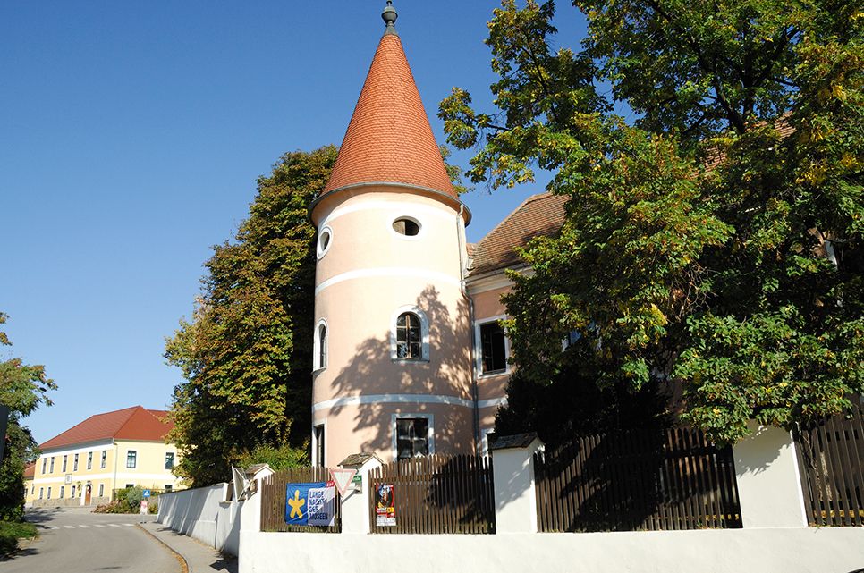 Ružová budova s vežou a červenou strechou vo Fels am Wagram, obklopená stromami a modrou oblohou.