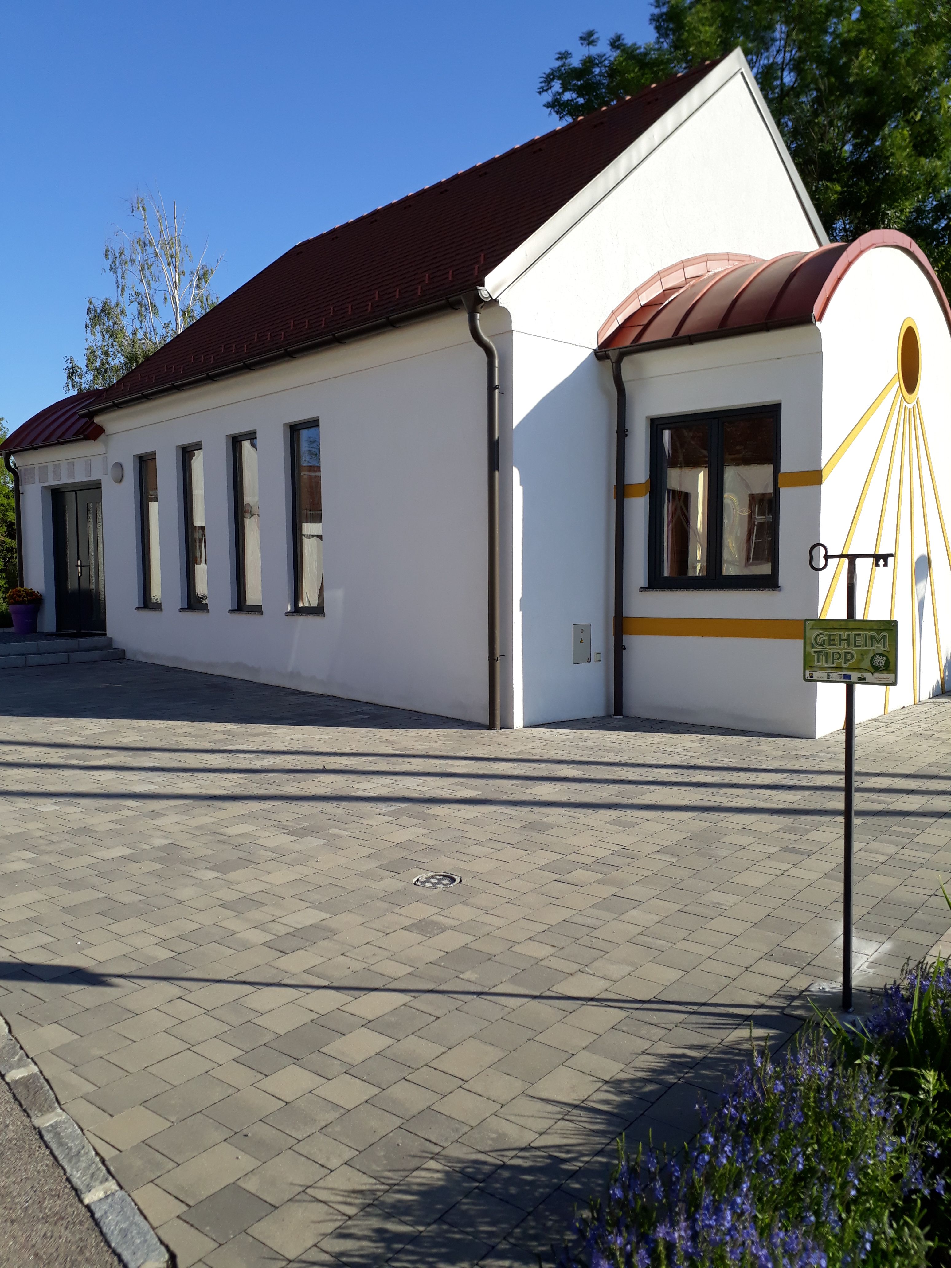 Biela kaplnka s červenou strechou a žltými akcentmi, obklopená dláždenou podlahou a rastlinami.