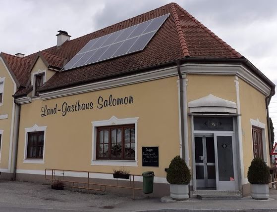 Pohľad zvonku na tradičný vidiecky hostinec so svetlou fasádou, červenými okenicami a šikmou strechou.