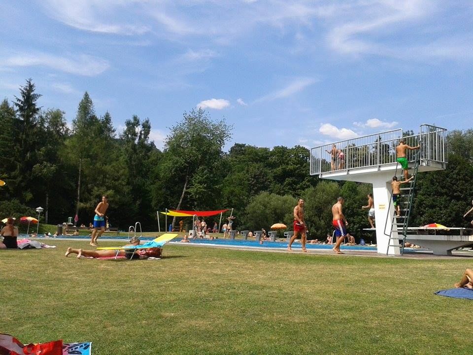 Ľudia v bazéne Seebensteinského parku, niektorí na trávniku, iní v bazéne. Vidno skokanskú vežu.