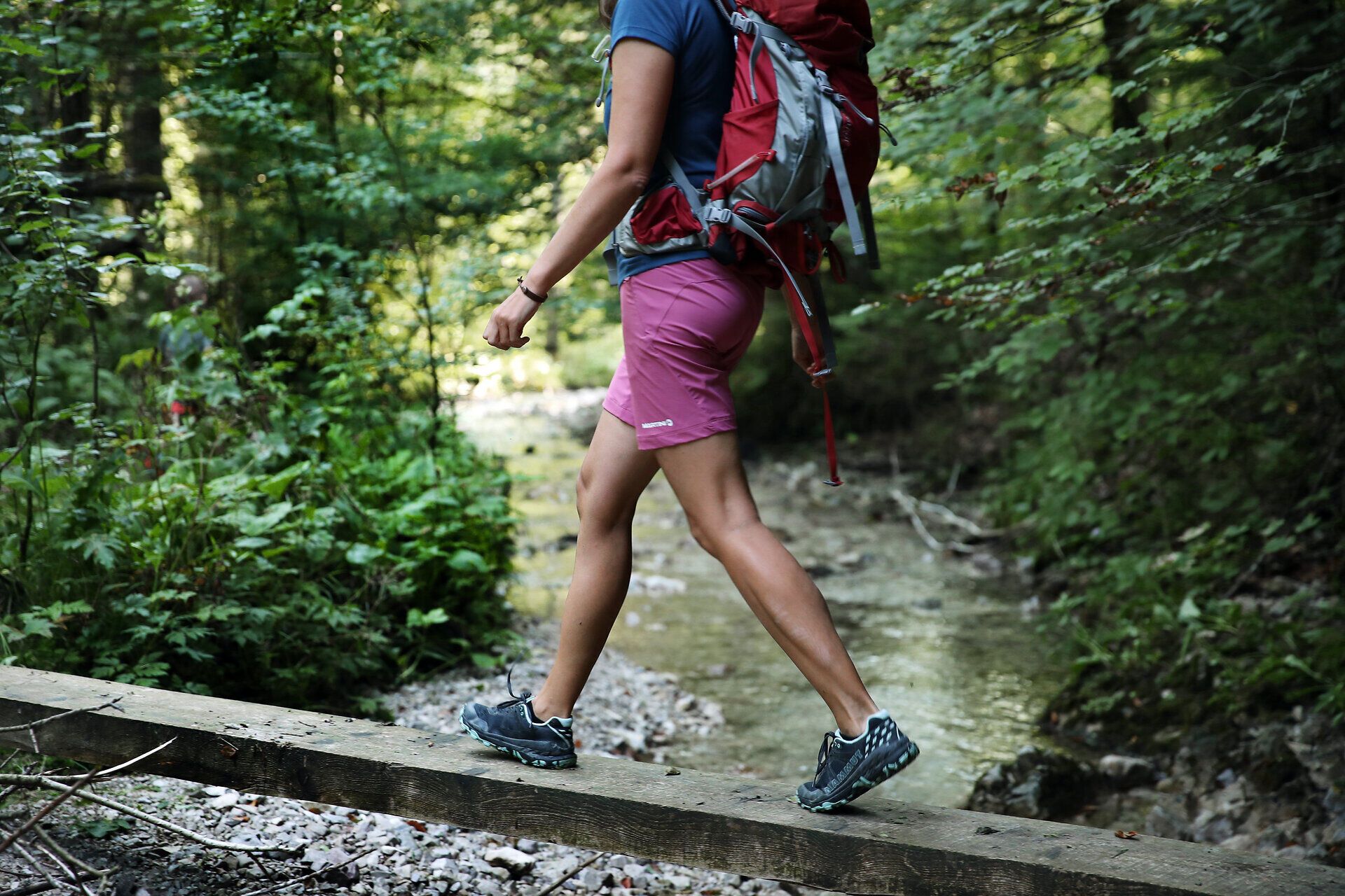 Žena v turistickom oblečení balansuje na drevených stĺpoch nad malým potokom. 