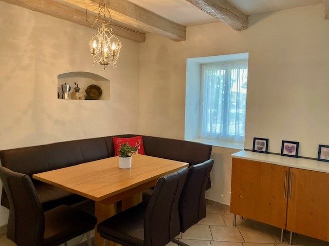 Útulný jedálenský kút s dreveným stolom, čiernymi stoličkami a rohovou lavicou. Na stene visí luster, vedľa neho je okno a príborník.