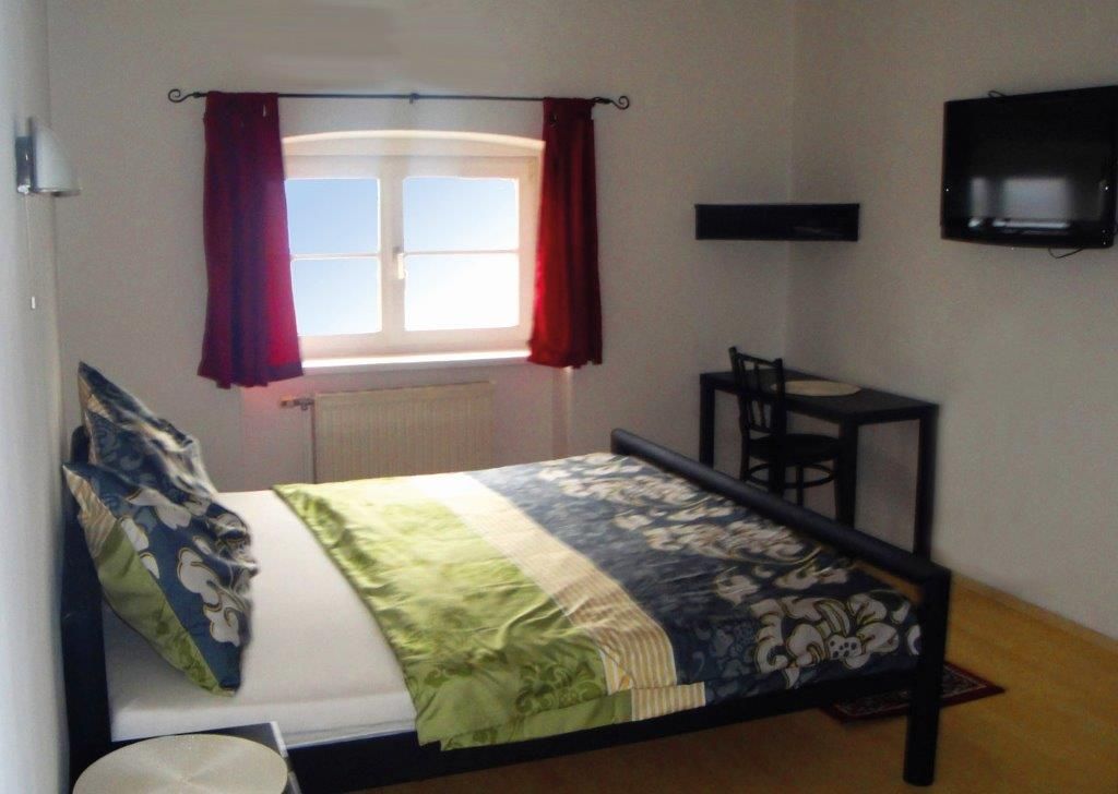 Spálňa s posteľou, oknom s červenými závesmi, malým stolom a stoličkou a televízorom na stene.