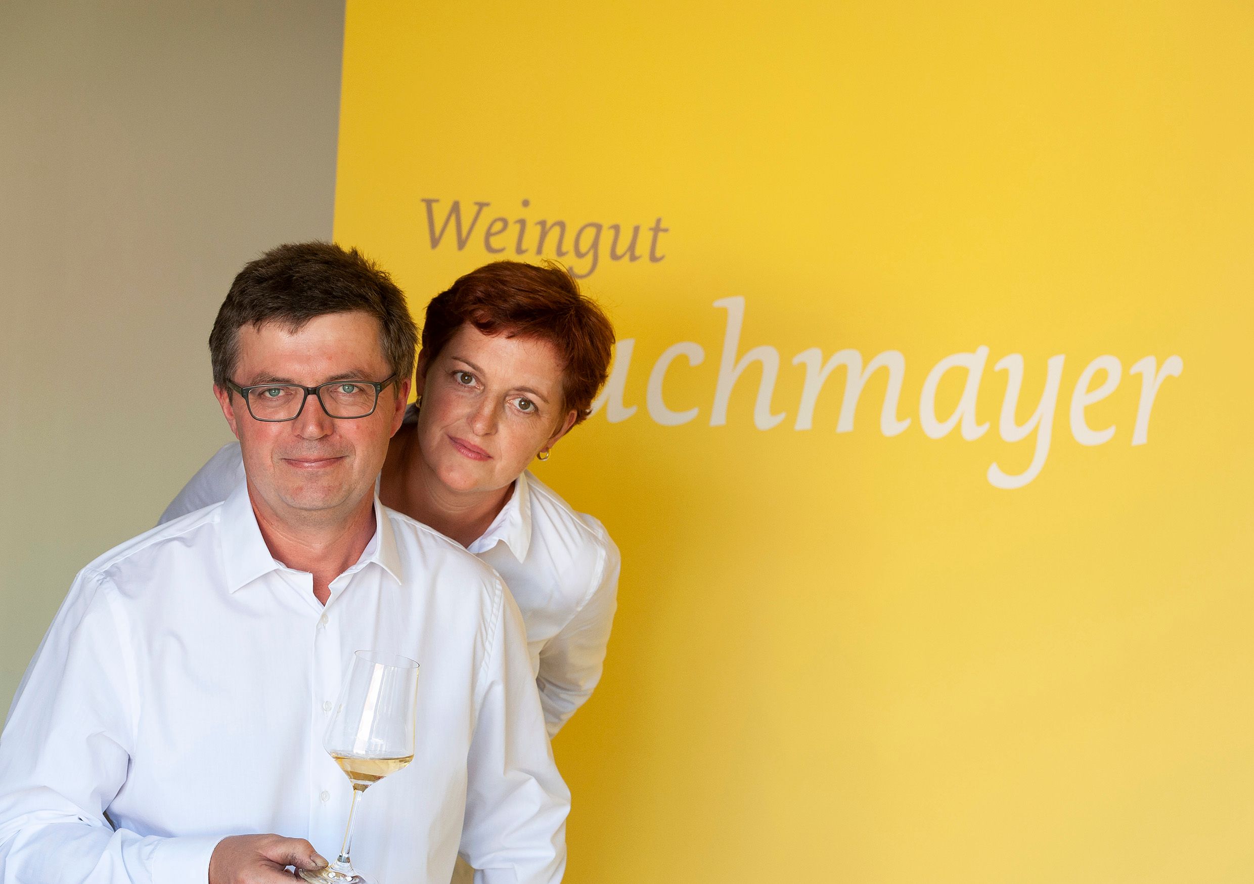Dve osoby v bielych košeliach na žltom pozadí s nápisom "Weingut Buchmayer". Jedna osoba drží pohár na víno.