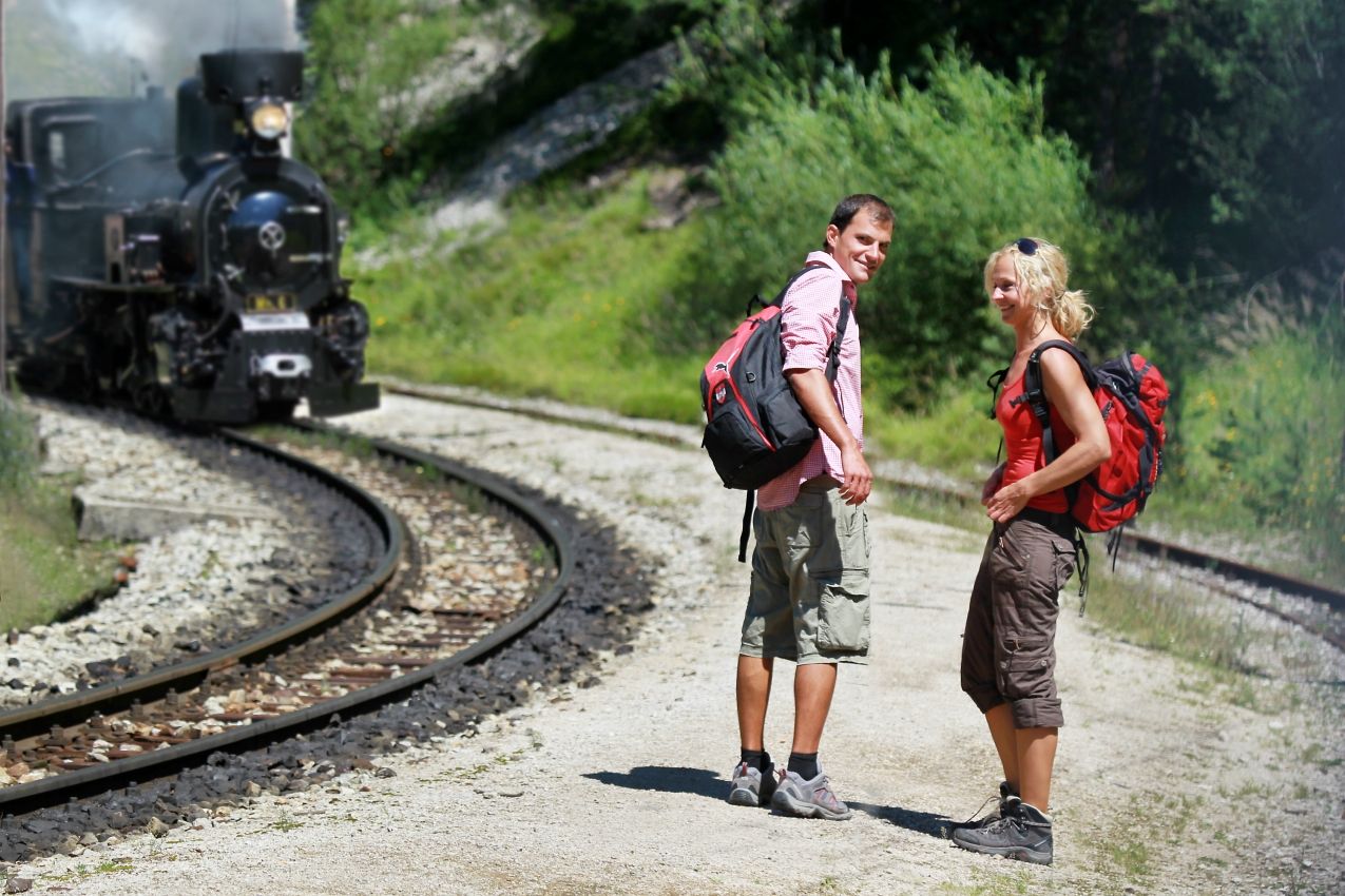Dvaja turisti stoja na štrkovej ceste vedľa železničných koľají a okolo nich prechádza parná lokomotíva.