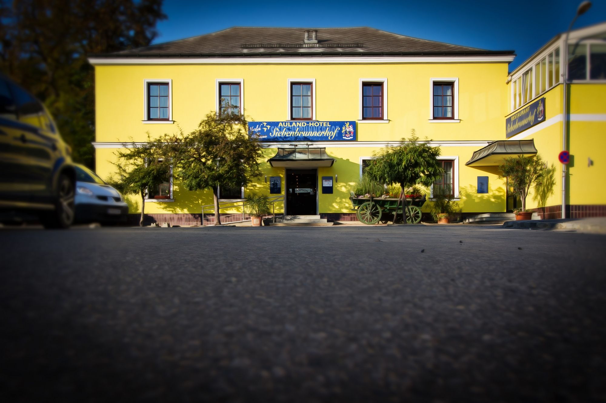 Žltá budova s nápisom "Auland-Hotel Siebenbrunnerhof", obklopená stromami a starým autom.
