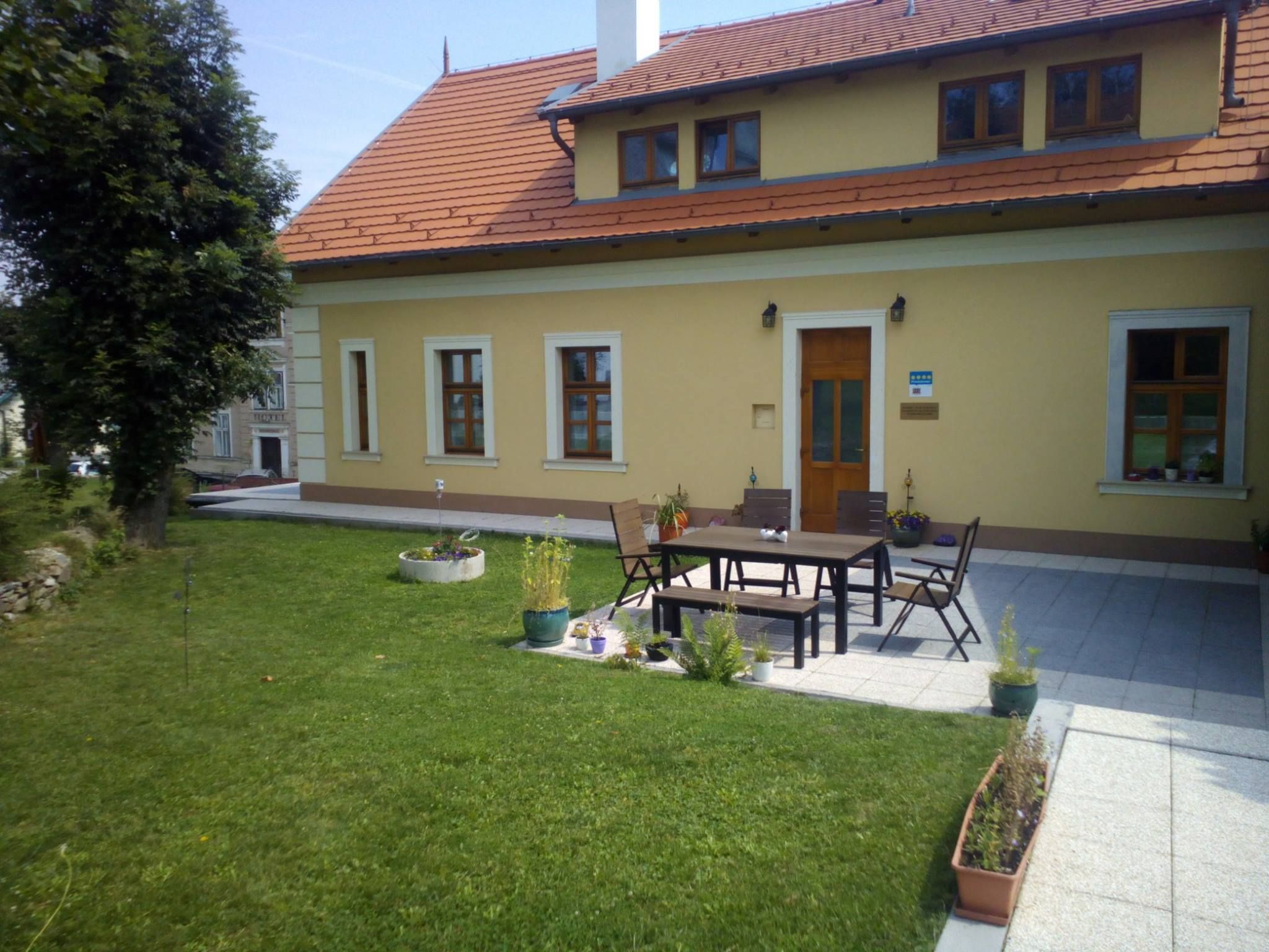 Žltý dom s červenou strechou a terasou, obklopený trávnikom a rastlinami.