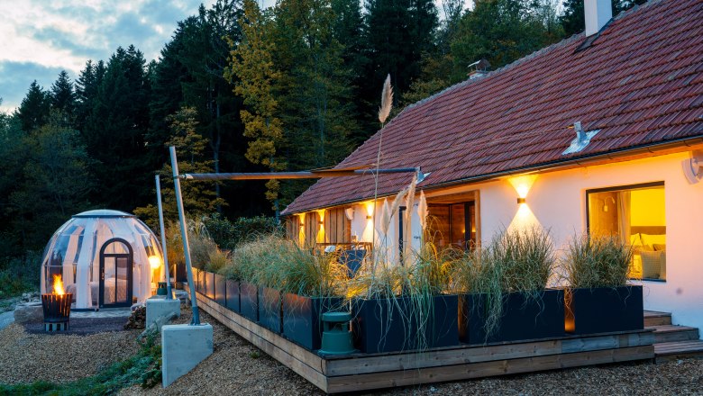 Chaty a miesta plné ZÁZRAKOV - Weidehaus De Luxe, © WUNDERVOLL eben Moderná chata s terasou a priehľadným kupolovitým stanom v lese za súmraku.