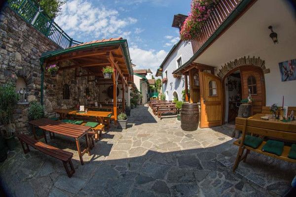 Útulné nádvorie s drevenými lavicami, stolmi a kvetmi. Kamenná podlaha a zastrešené miesta na sedenie. Slnečné počasie s modrou oblohou.