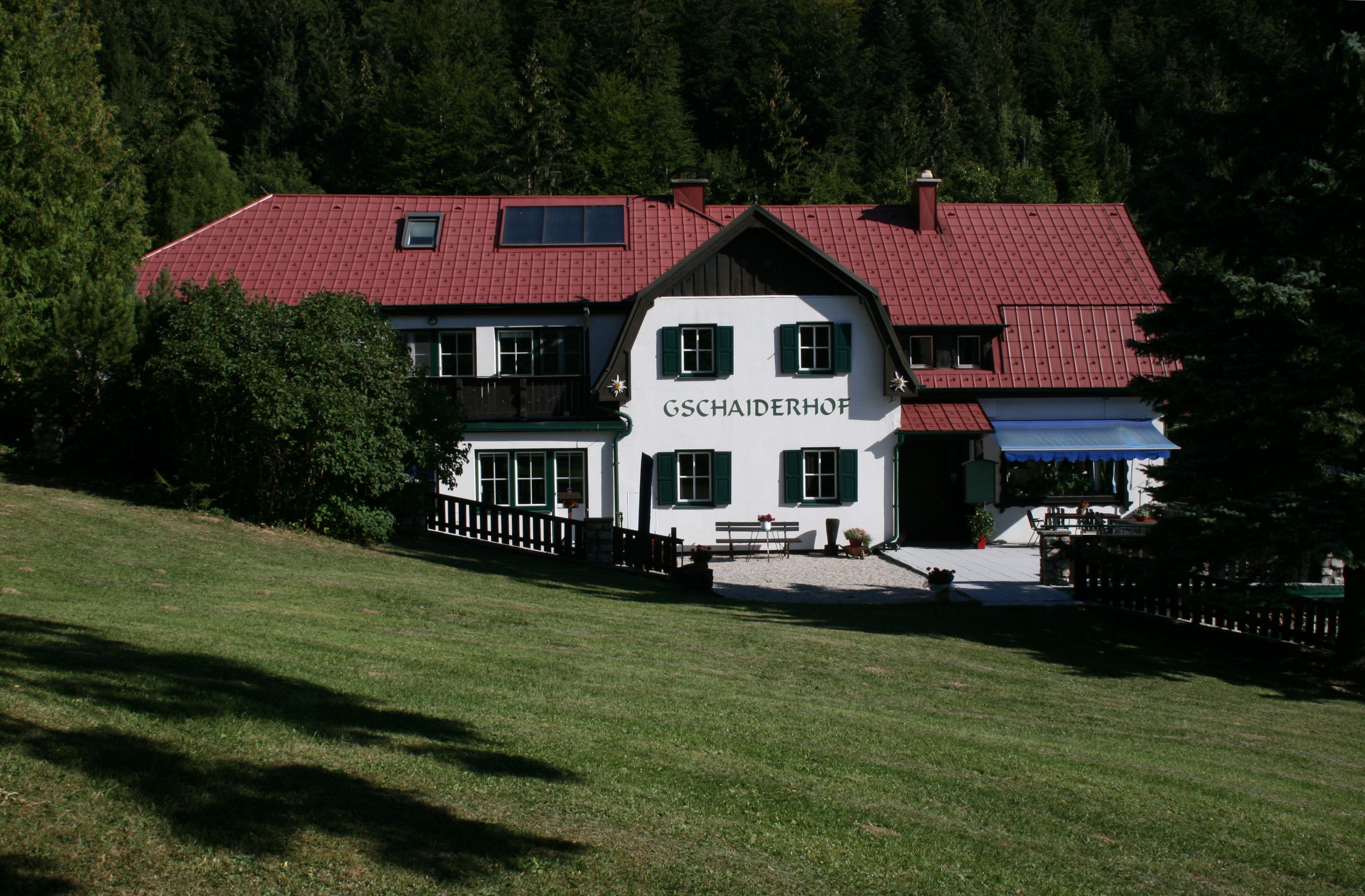 Tradičná budova s červenou strechou a zelenými okenicami, obklopená stromami a lúkami, s nápisom "Gschaiderhof".