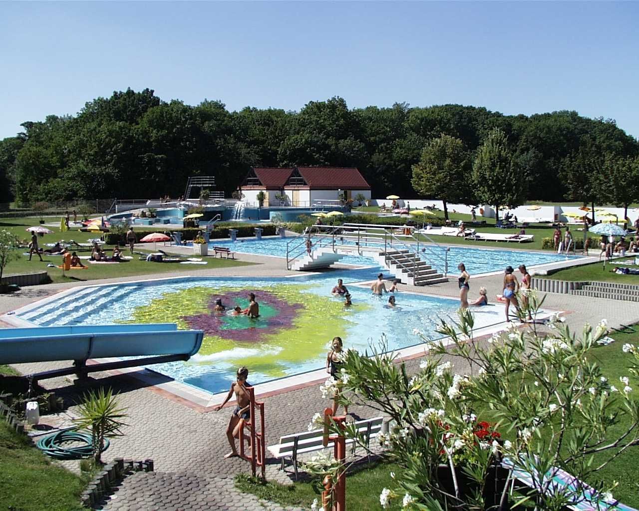 Vonkajší bazén s niekoľkými bazénmi, šmykľavkou a trávnikom na opaľovanie, obklopený stromami.