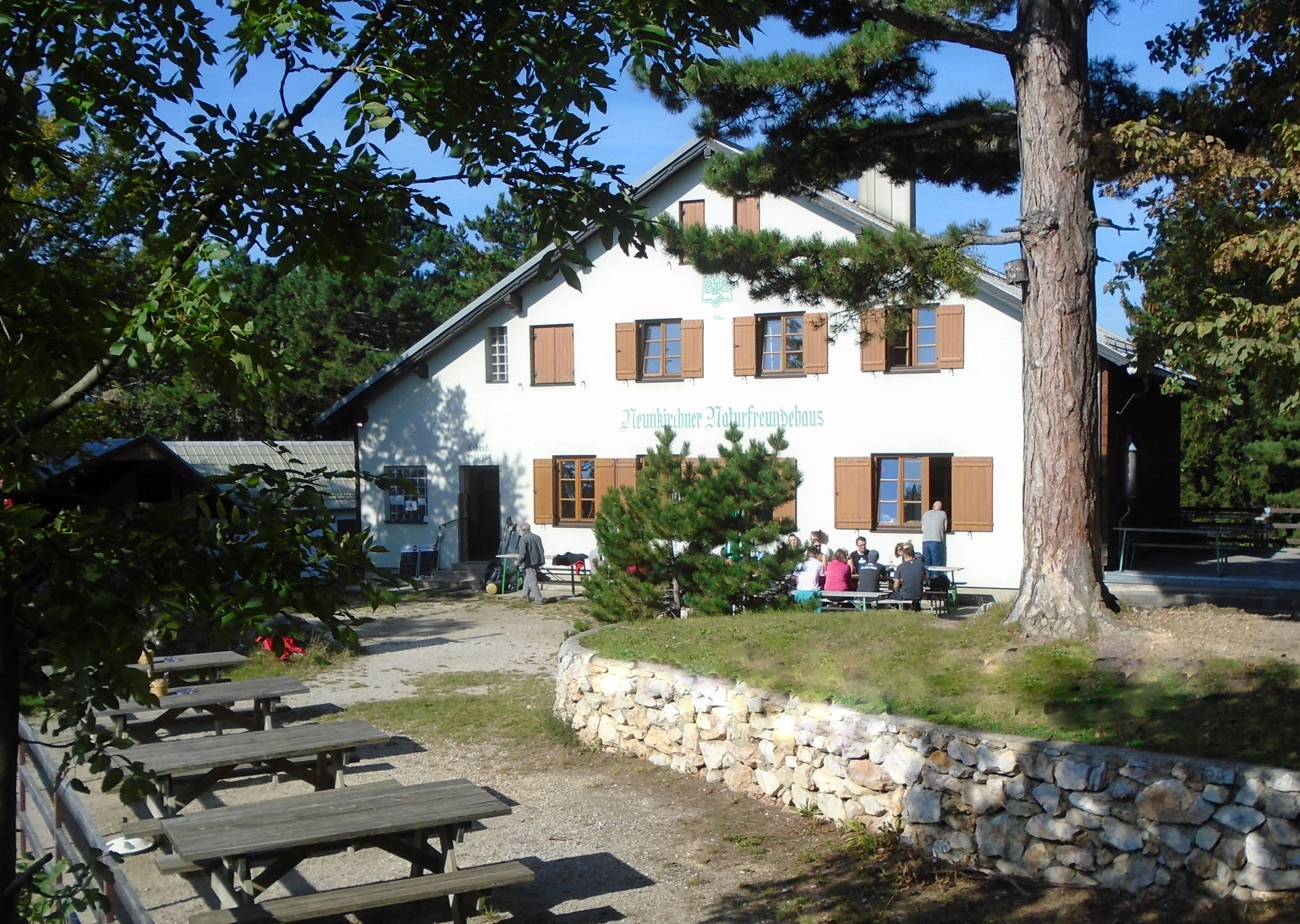 Budova s nápisom "Neunkirchner Naturfreundehaus", vonku obklopená stromami a lavičkami.