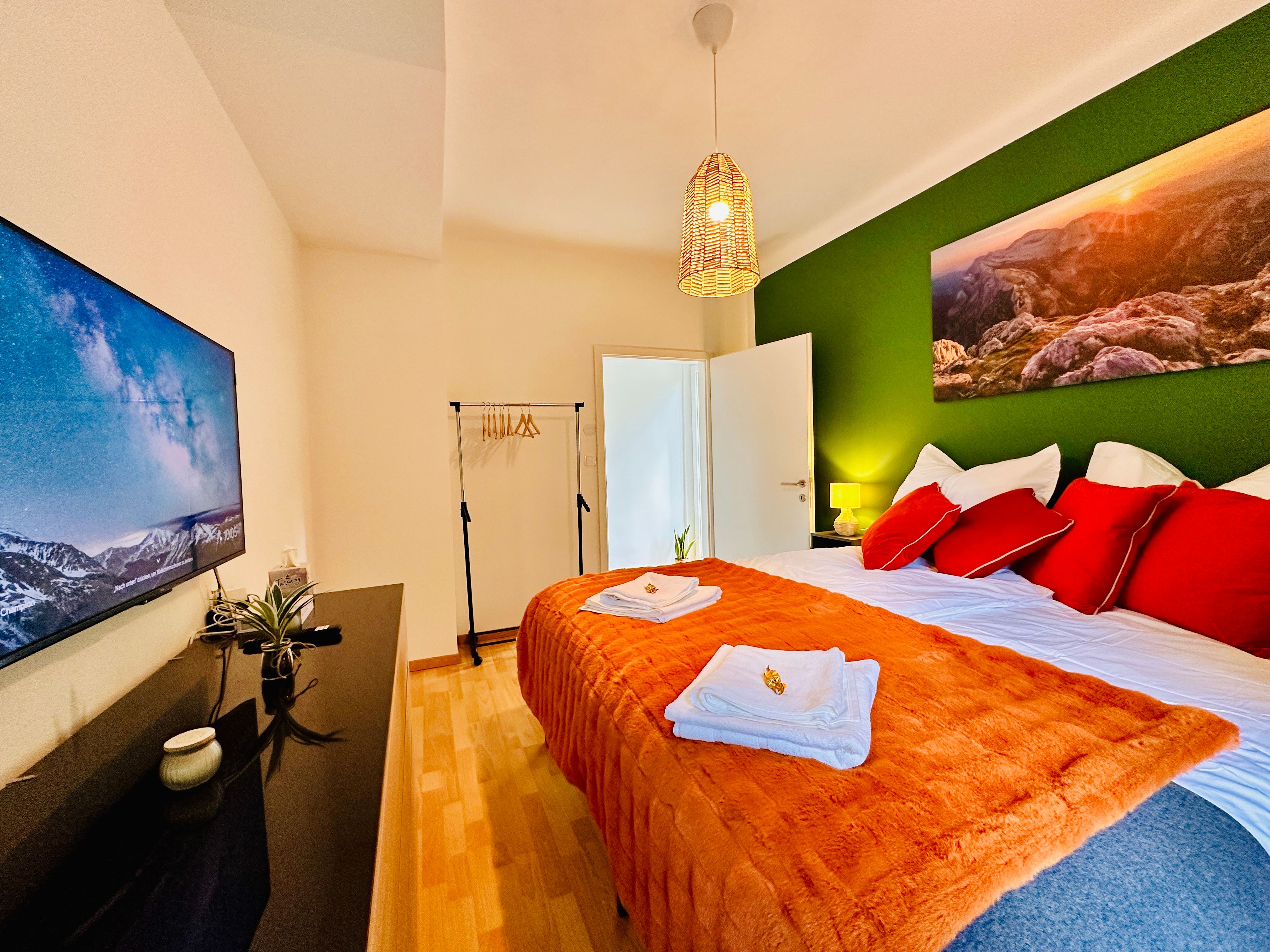 Útulná spálňa so zeleným akcentom, manželskou posteľou, TV a dekoráciou.