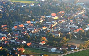 Letecký pohľad na dedinu s kostolom a domami, obklopenú stromami a poľami.