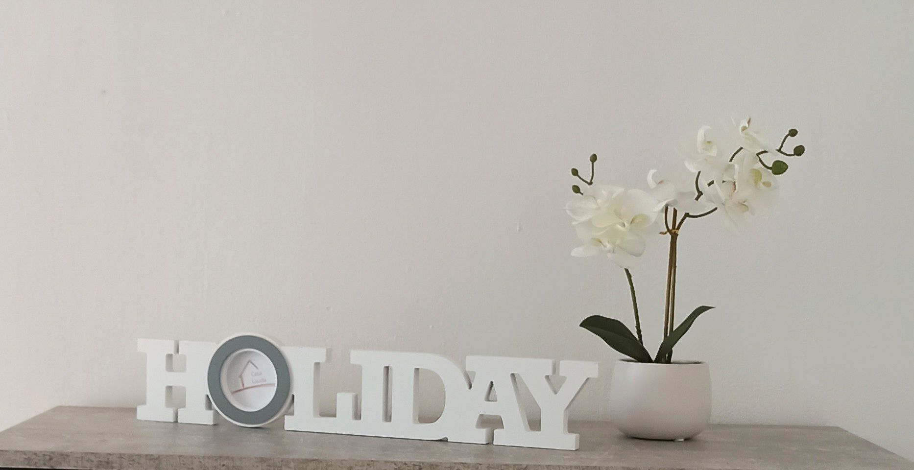 Dekorácia s nápisom "HOLIDAY" a bielou orchideou v kvetináči na stole.