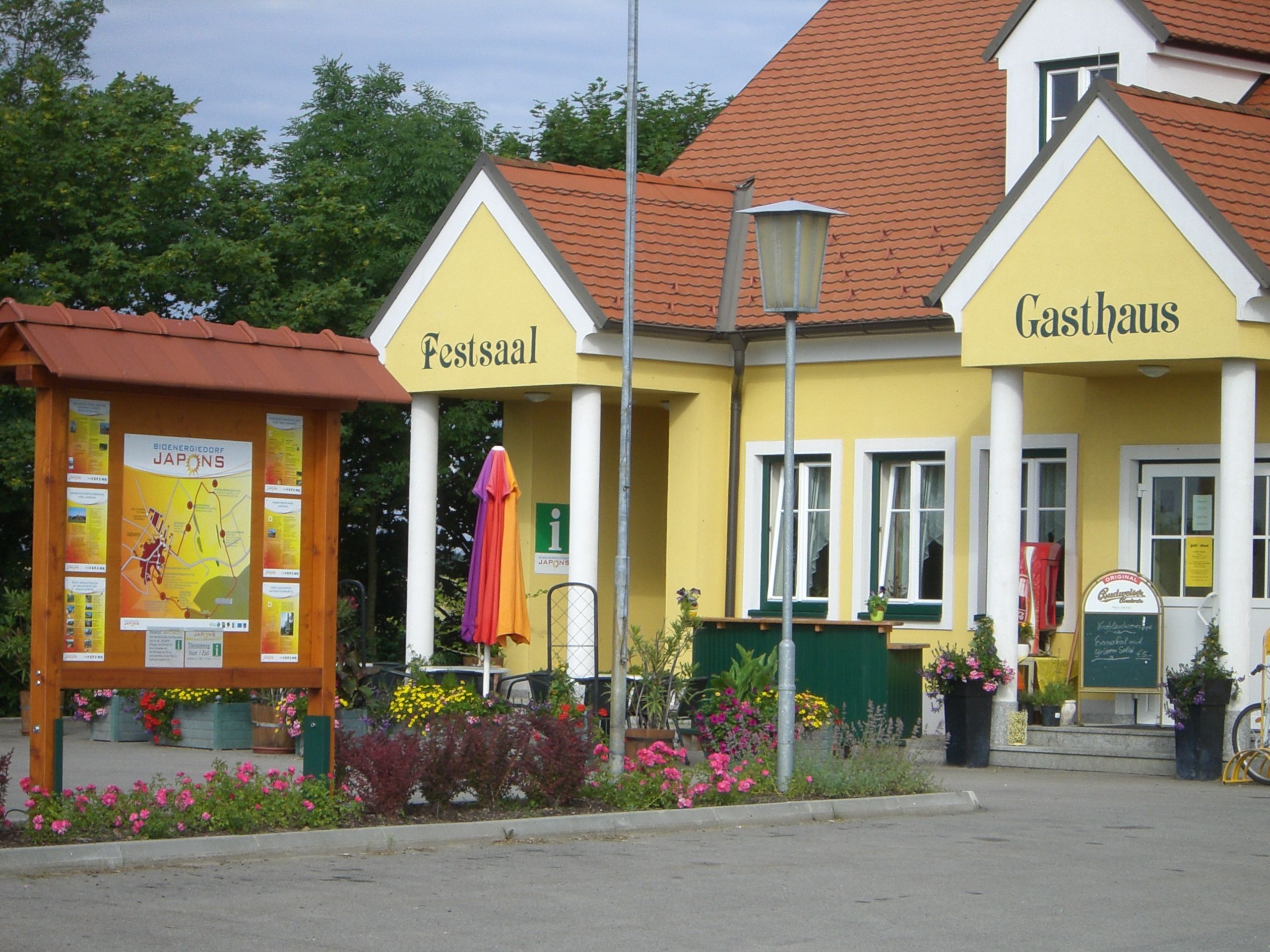 Žltá budova s nápismi "Festsaal" a "Gasthaus", obklopená kvetmi a informačnou tabuľou.