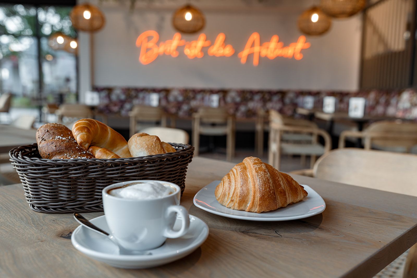 Kaviareň s košíkom pečiva, croissantom na tanieri a šálkou cappuccina na stole.
