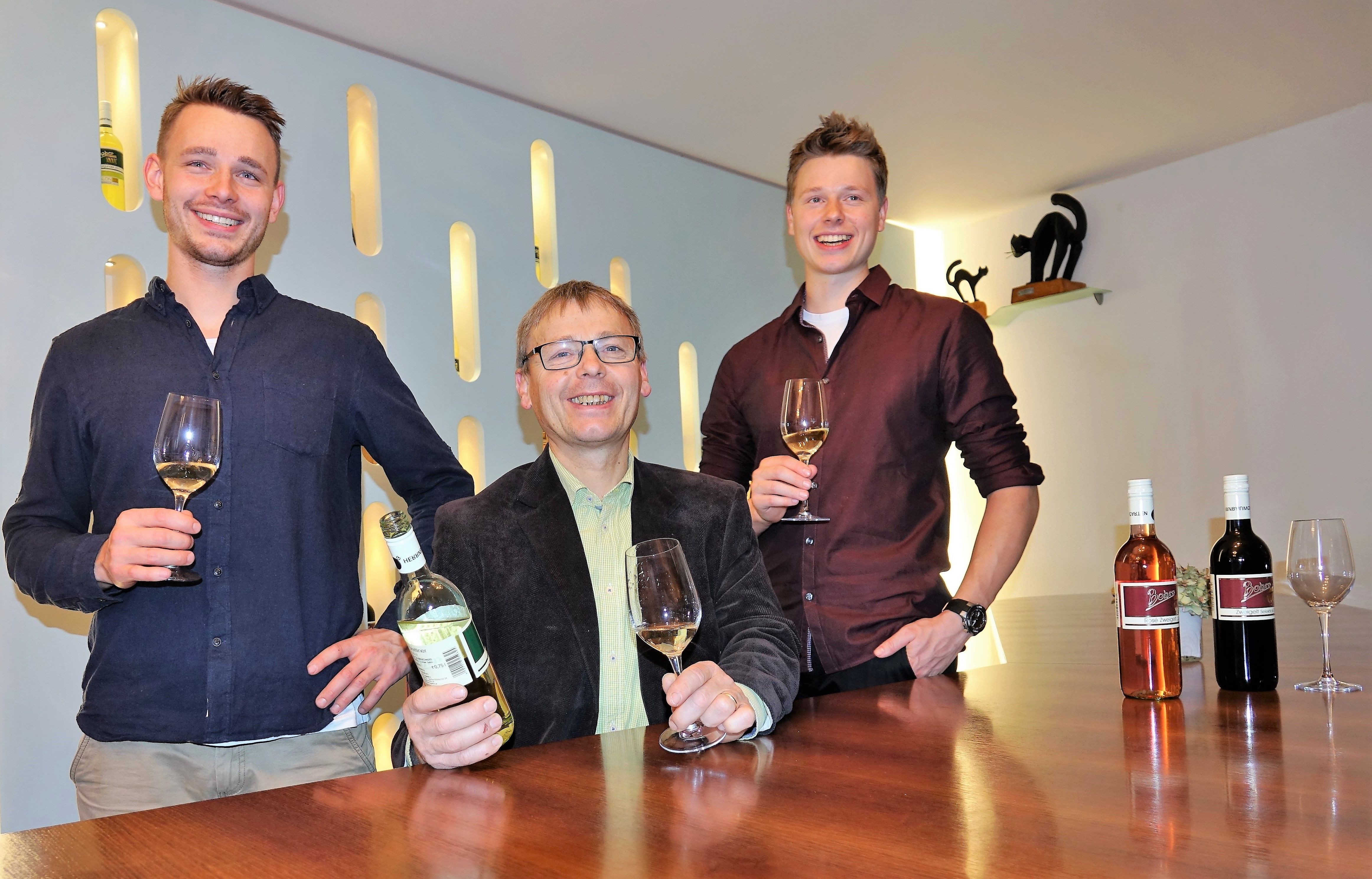 Traja muži stoja pri stole s pohármi a fľašami vína.