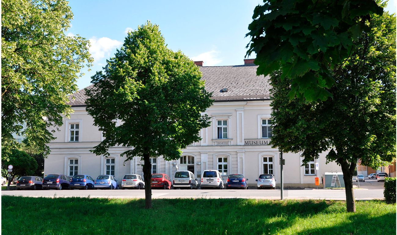 Biela budova s nápisom "Múzeum", obklopená stromami a zaparkovanými autami.
