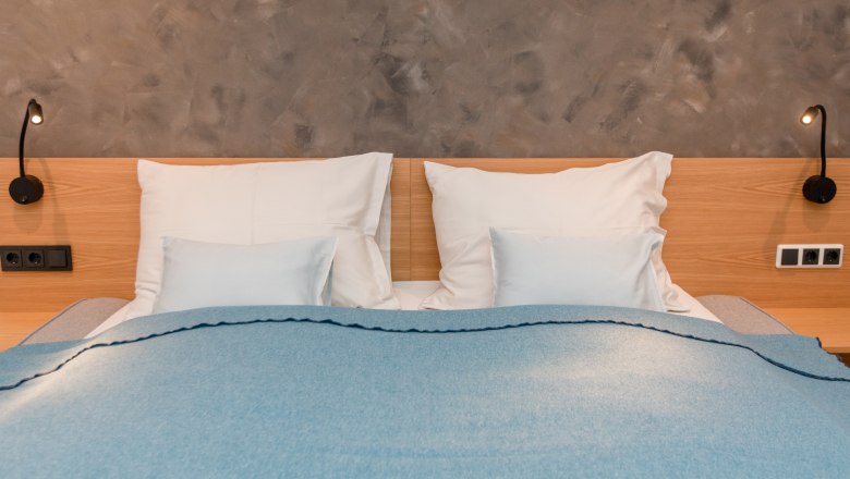 Manželská posteľ s modrou prikrývkou a bielymi vankúšmi pred sivou stenou.
