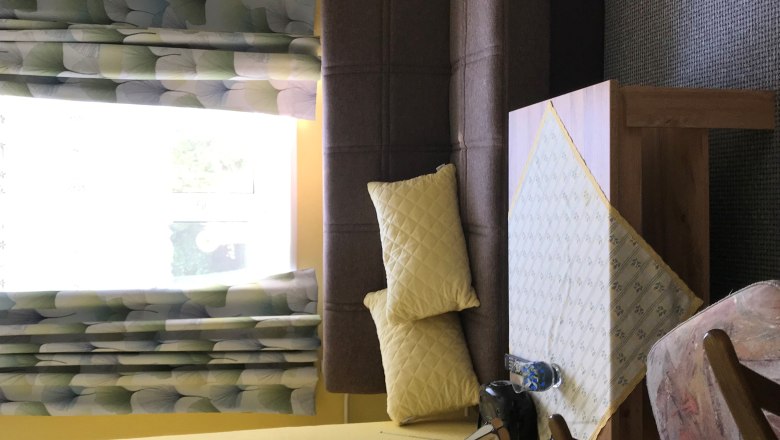 Ob&yacute;vacia izba s hnedou pohovkou, žlt&yacute;mi vank&uacute;&scaron;mi, stolom s obrusom, kreslom a oknom so vzorovan&yacute;mi z&aacute;vesmi.