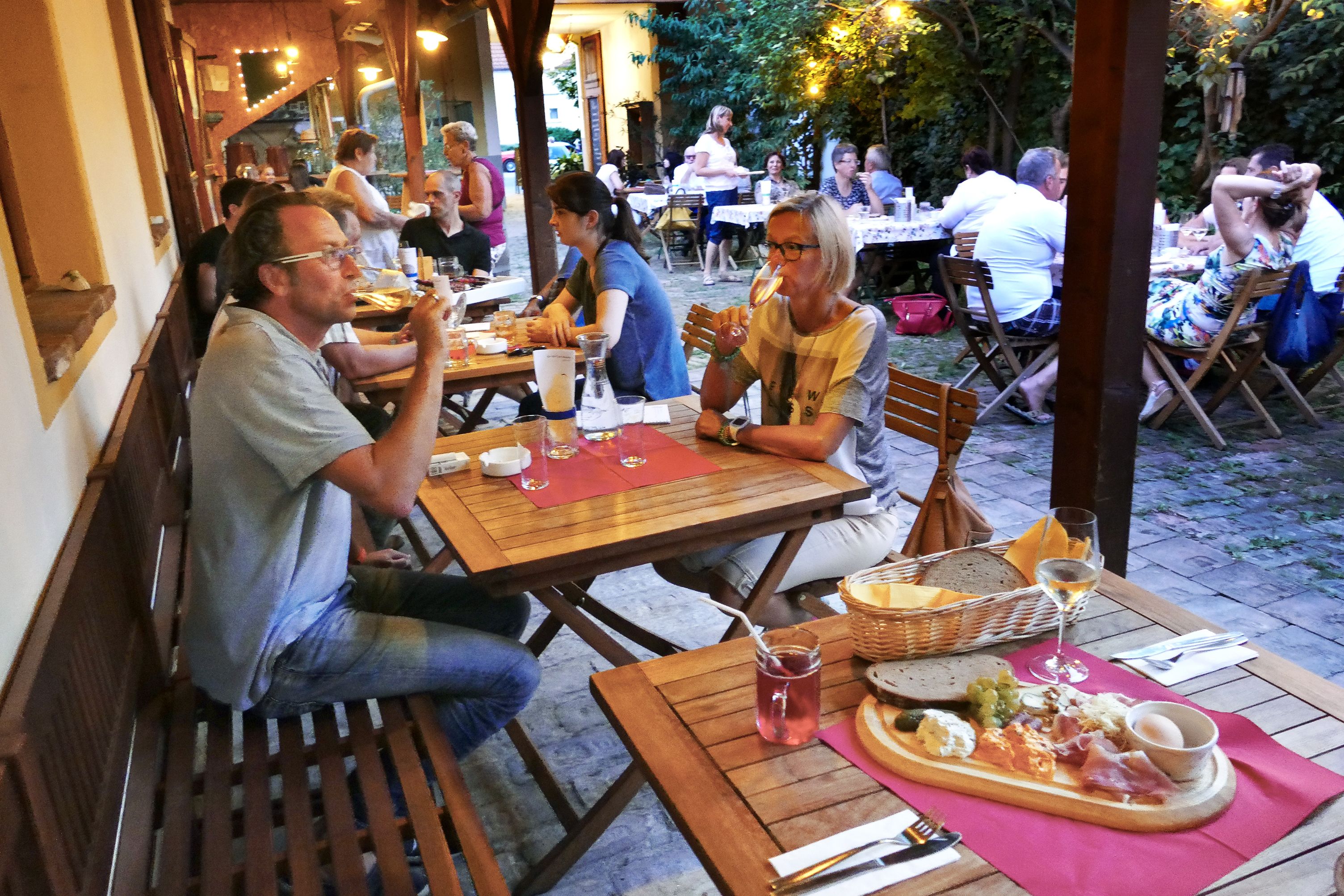 Ľudia sedia pri drevených stoloch v útulnom vonkajšom priestore a vychutnávajú si jedlo a nápoje.