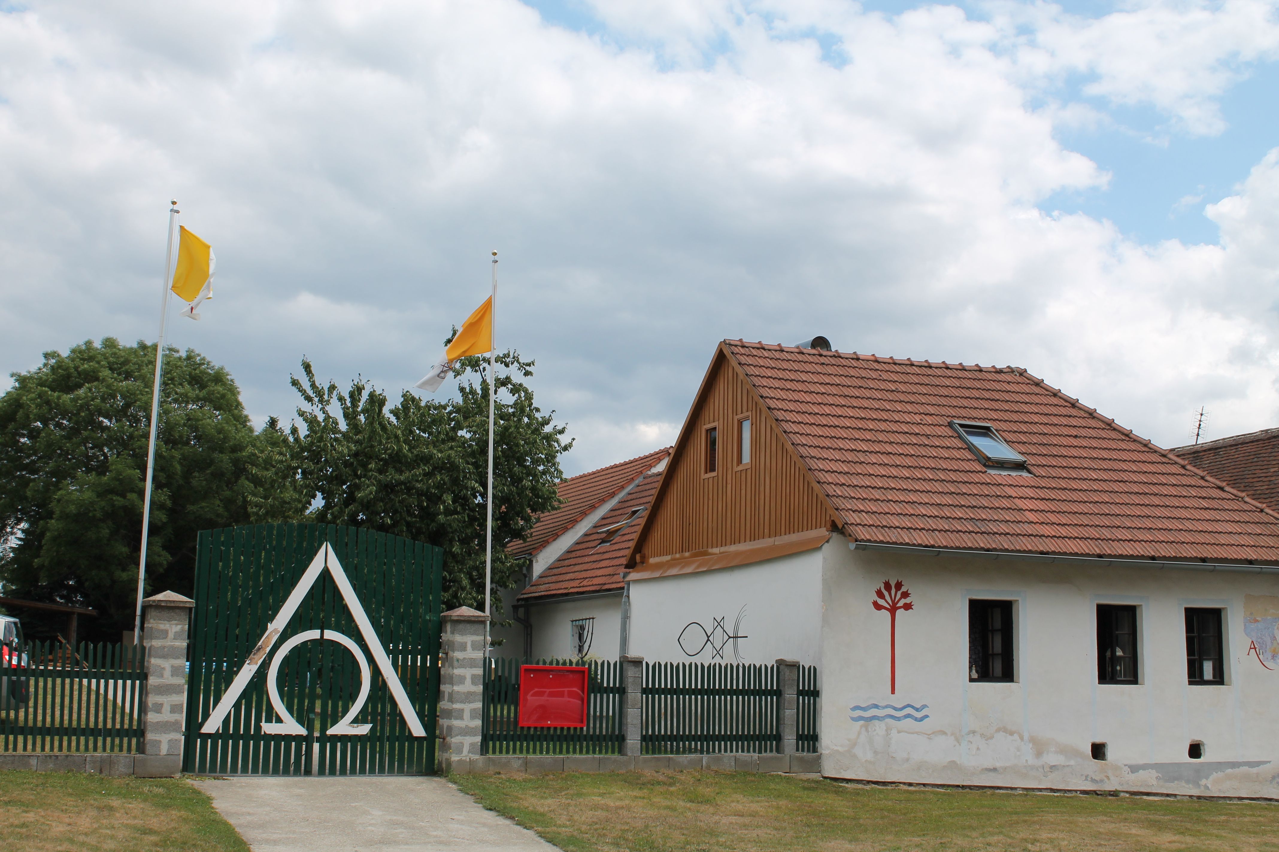 Dom s červenými škridlami, zelený plot so symbolom Alfa Omega, dve žlté vlajky a nástenné maľby.