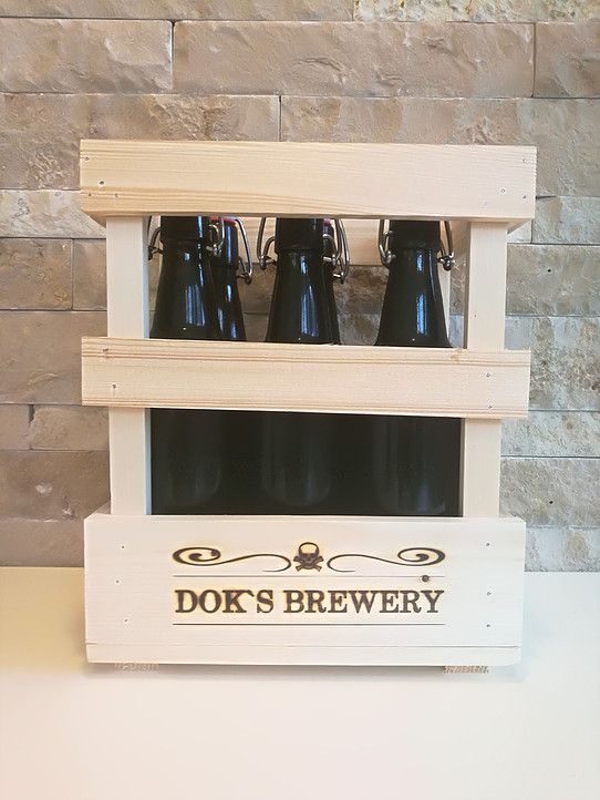 Drevená debna s tromi tmavými fľašami piva z pivovaru Dok's Brewery pred kamennou stenou.