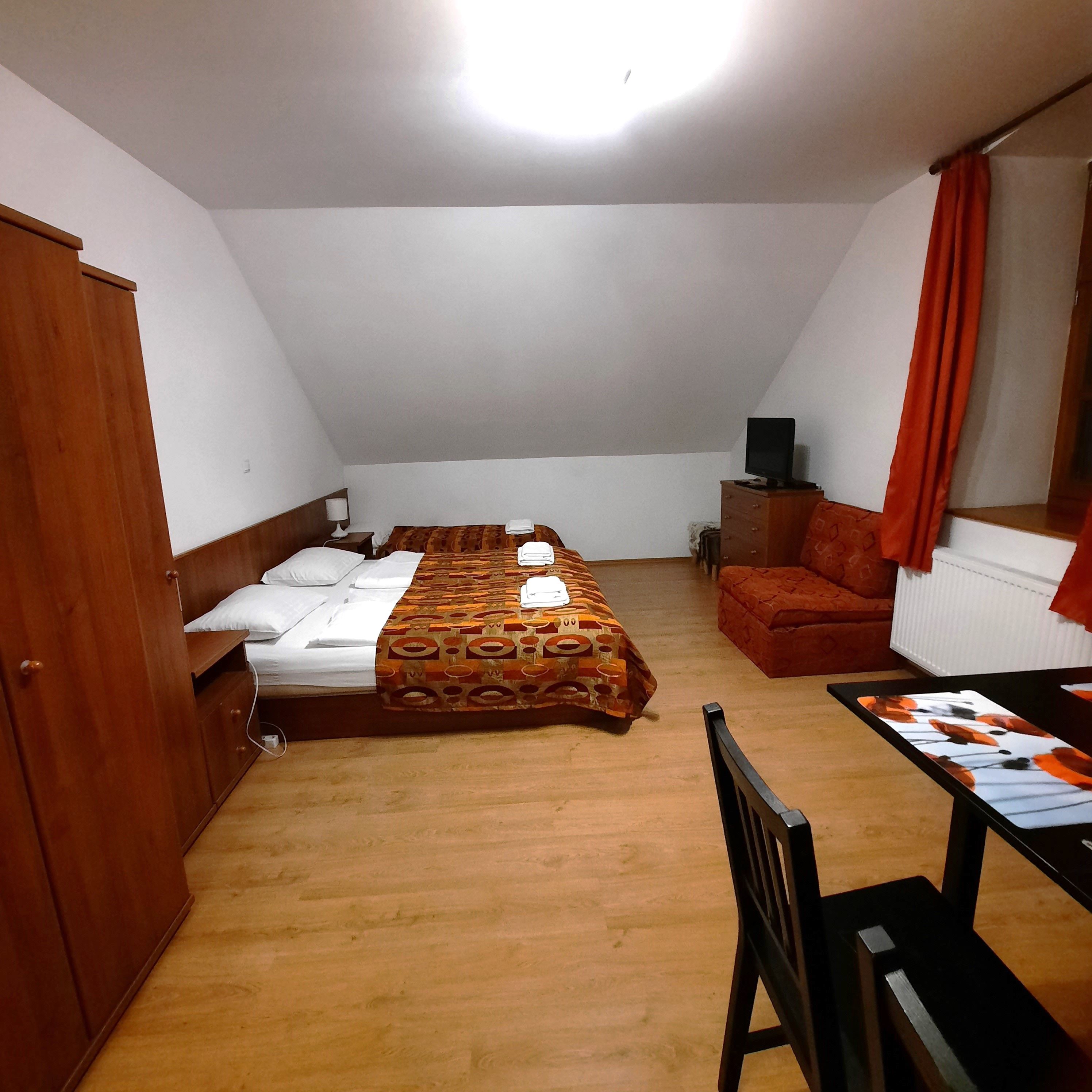 Jednoducho zariadená izba s manželskou posteľou, pohovkou, stolom a stoličkou. V miestnosti dominuje drevený nábytok a závesy oranžovej farby.