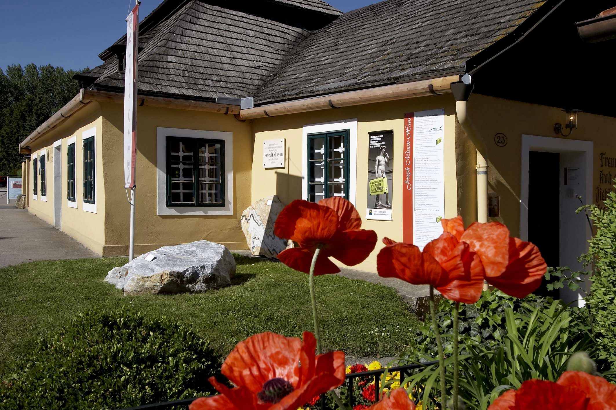 Žltý dom so šindľovou strechou a makmi v popredí.