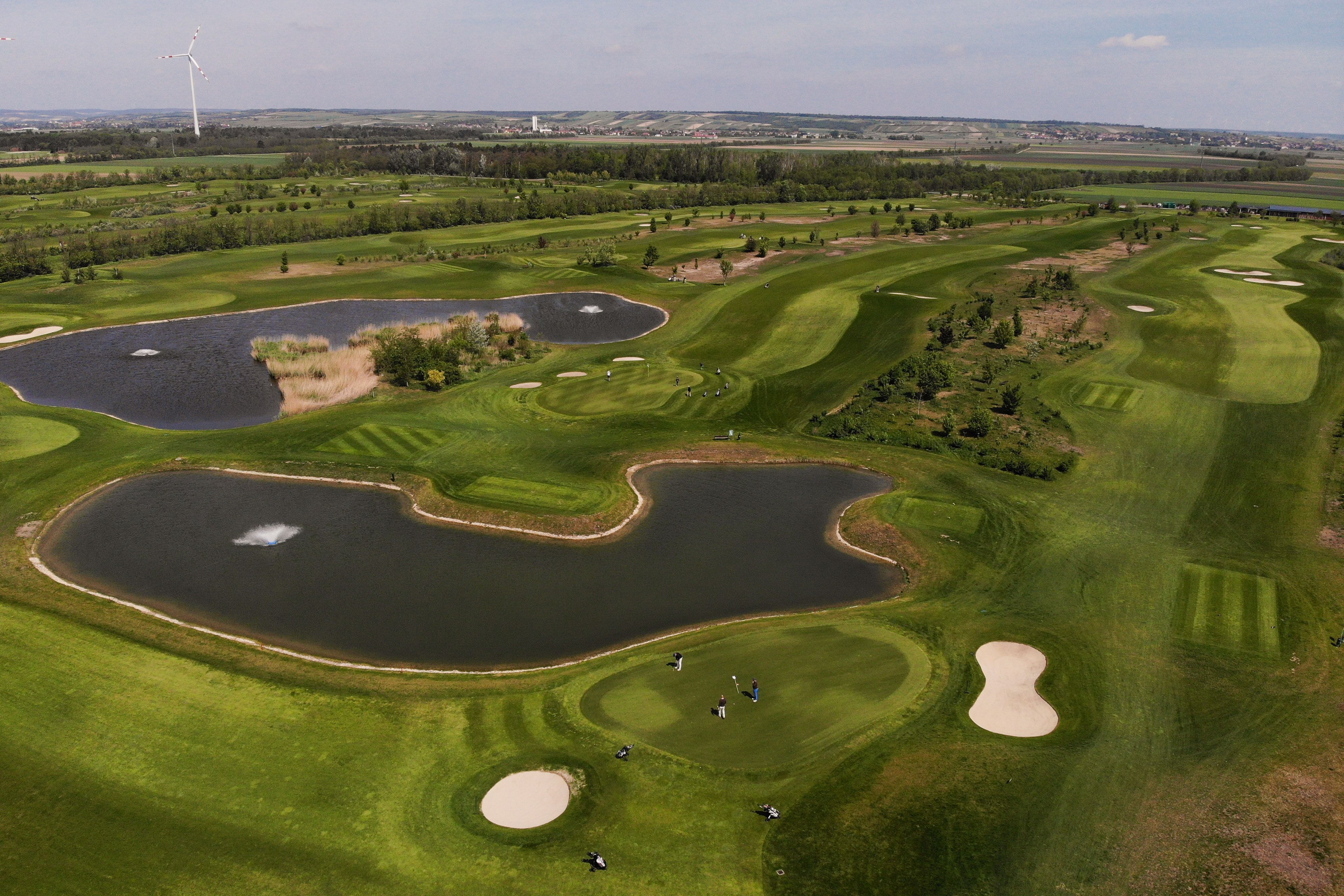 Letecký pohľad na golfové ihrisko s rybníkmi a veternou turbínou v pozadí.