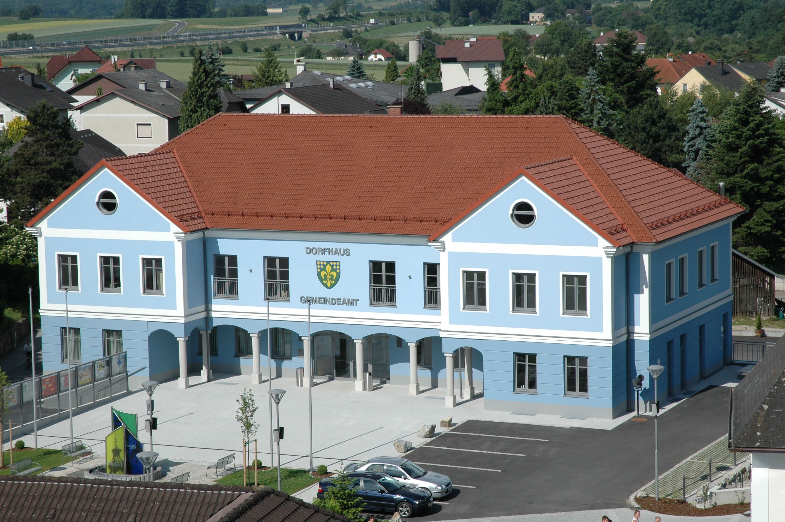 Modrá budova s červenou strechou, označená ako obecný úrad, obklopená domami a stromami.