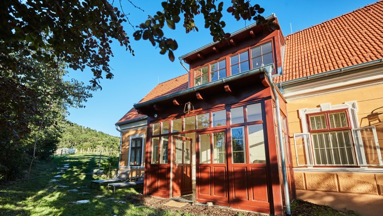 Žltá vila s červenými škridlami a preskleným vchodom, obklopená stromami a zelenou záhradou.