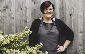 Žena v okuliaroch a zástere stojí s úsmevom pred drevenou stenou vedľa kríka.