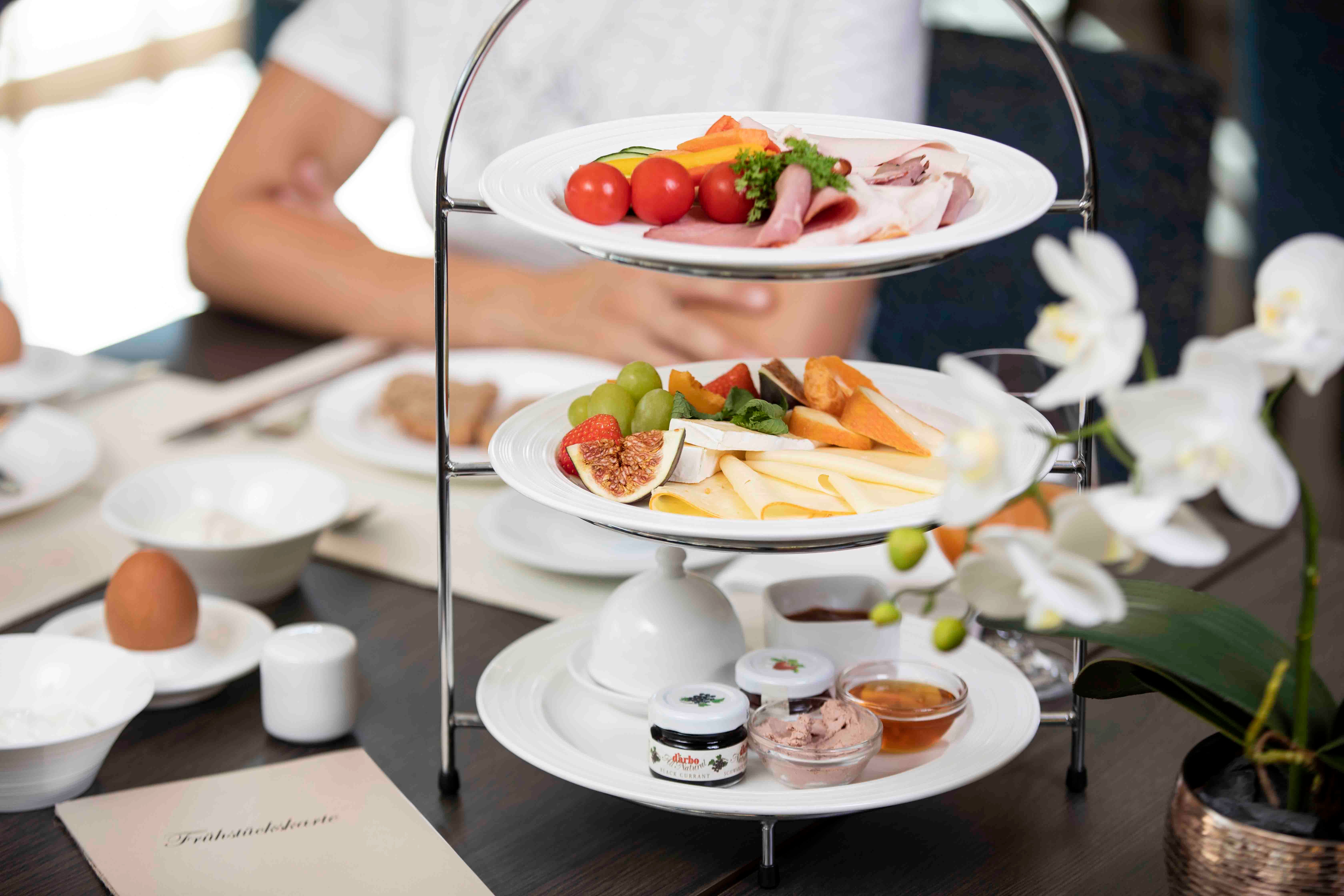 Raňajkový stôl s etážou plnou syrov, údenín, ovocia a nátierok. V popredí biela orchidea.