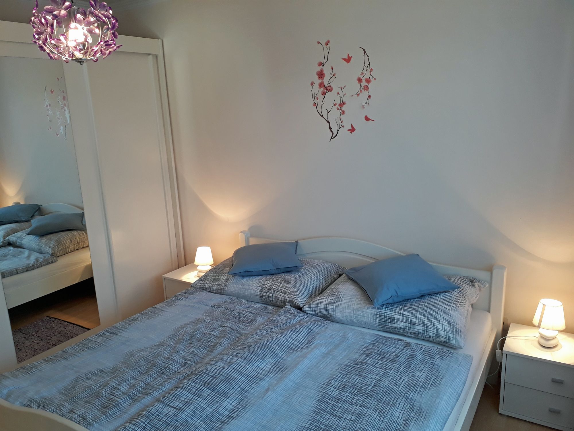 Útulná spálňa s manželskou posteľou, modrou posteľnou bielizňou, nočnými stolíkmi s lampami a nástenným tetovaním s kvetmi a vtákmi.