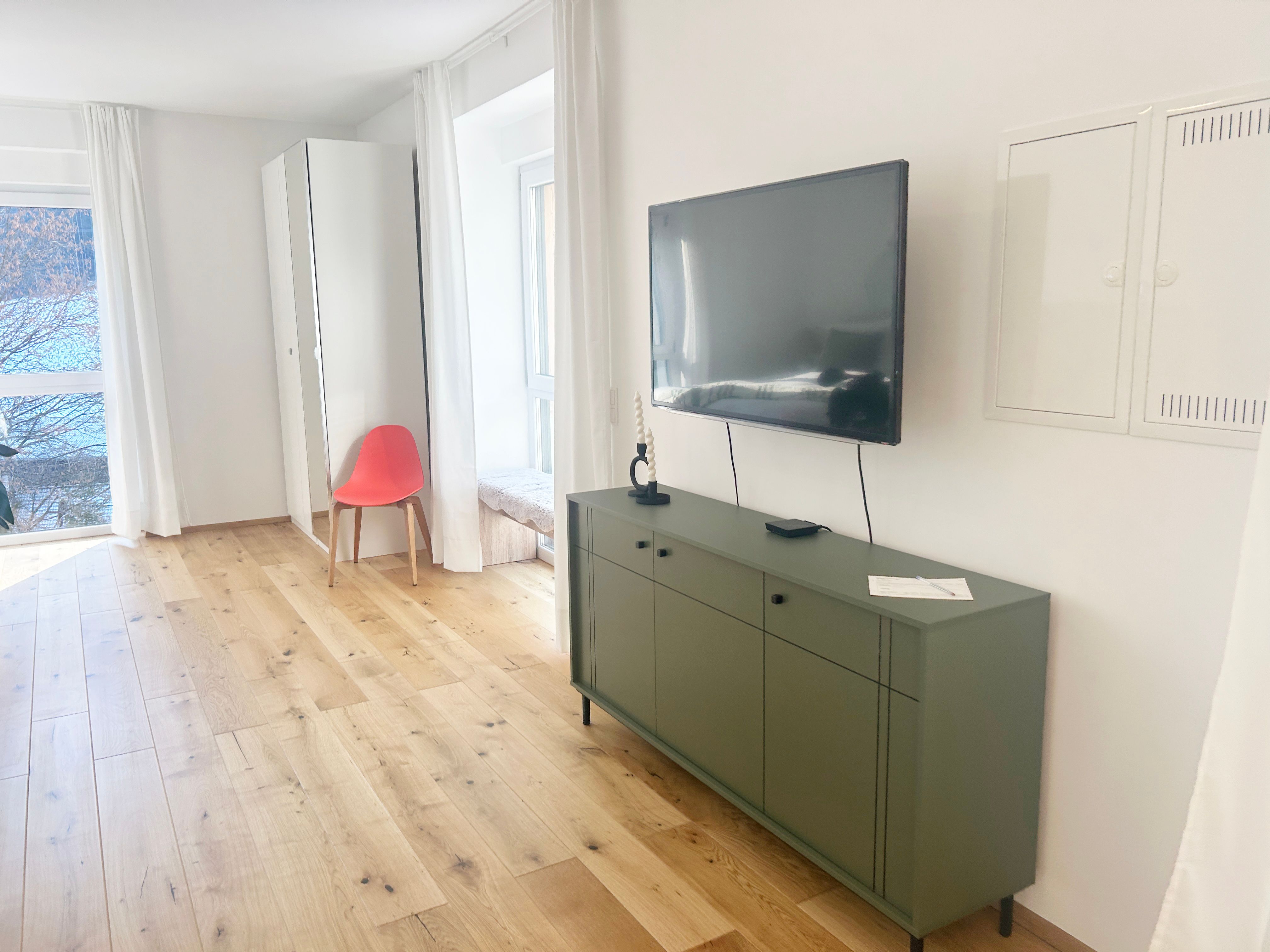 Moderná obývacia izba s drevenou podlahou, zeleným príborníkom, nástenným televízorom a červeným kreslom pred zrkadlom.
