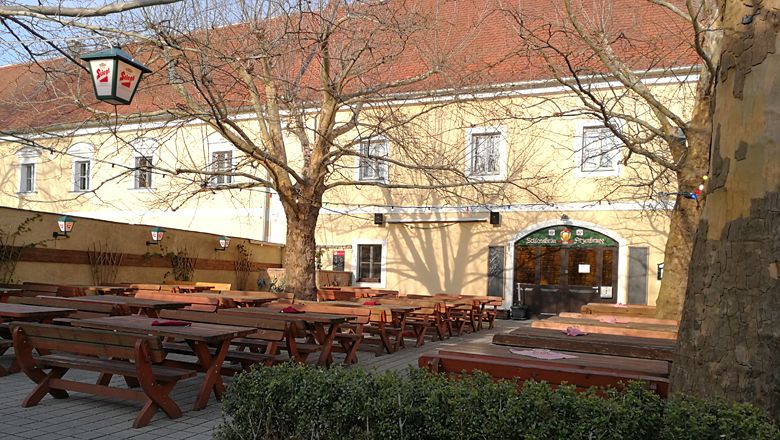 Pivná záhrada s drevenými lavicami a stolmi pred žltou budovou.