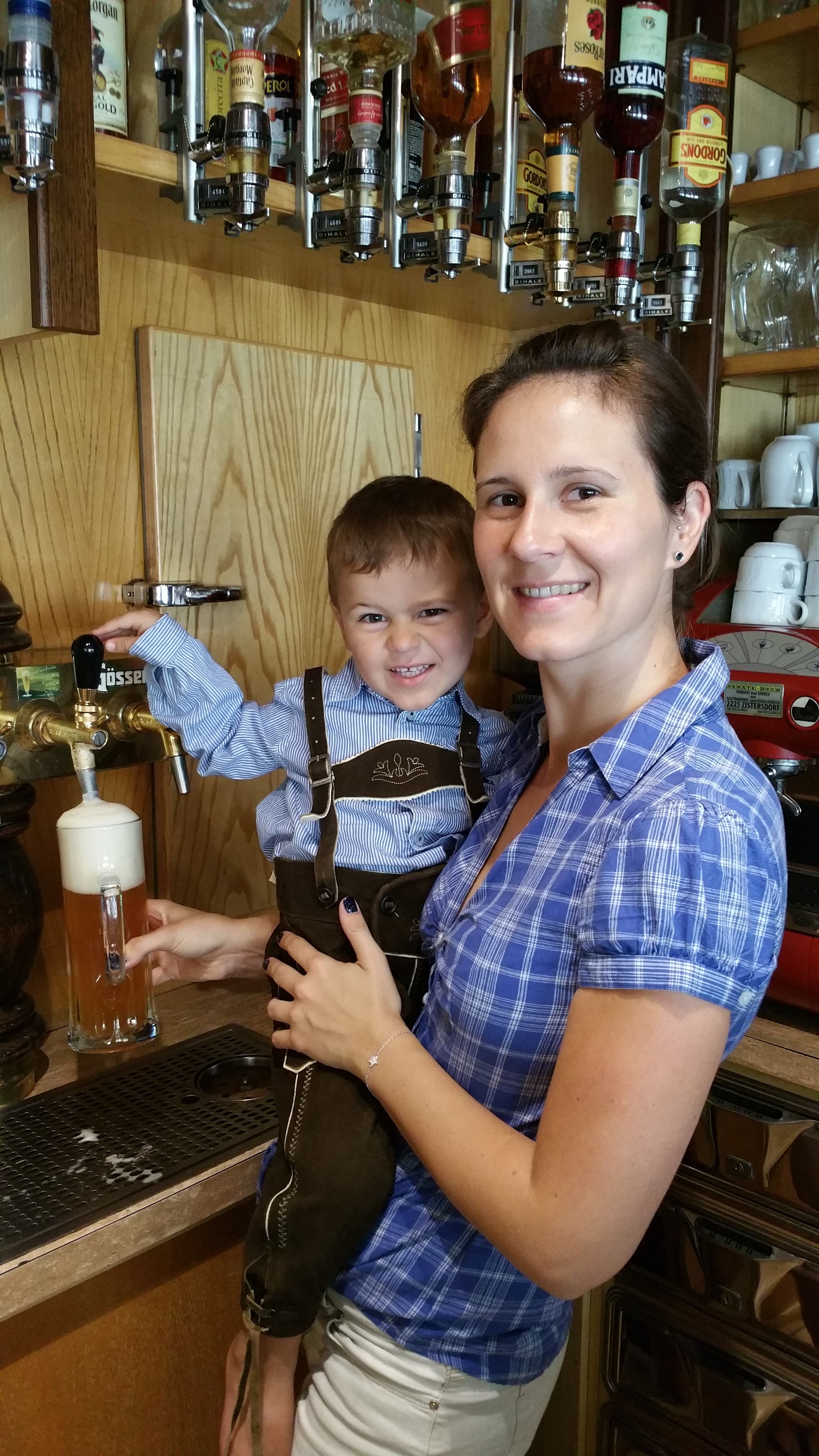 Žena drží dieťa v lederhosen pri výčape piva. V pozadí vidno fľaše s liehovinami.