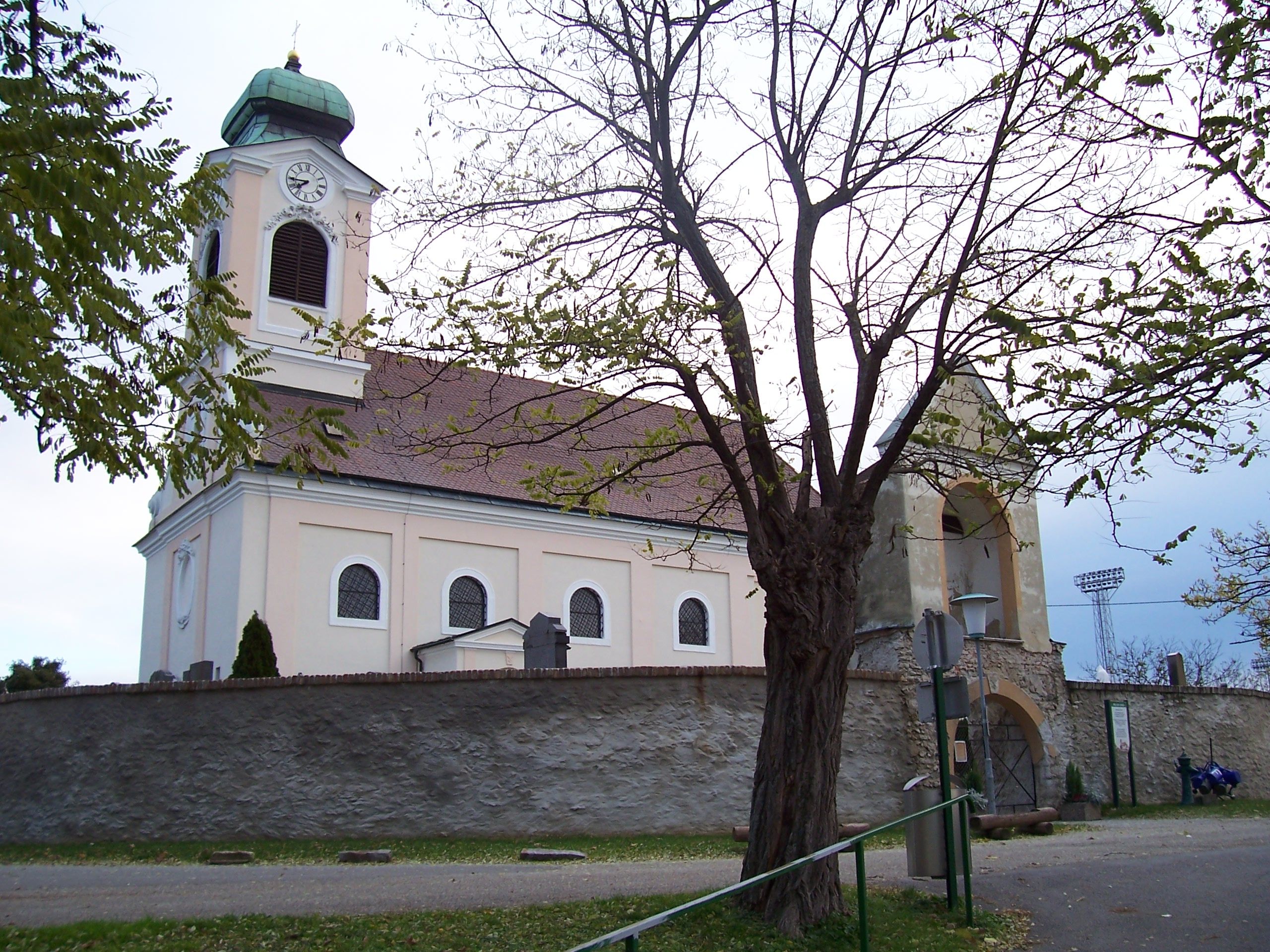 Kostol s vežou a hodinami, obklopený stromami a kamenným múrom.