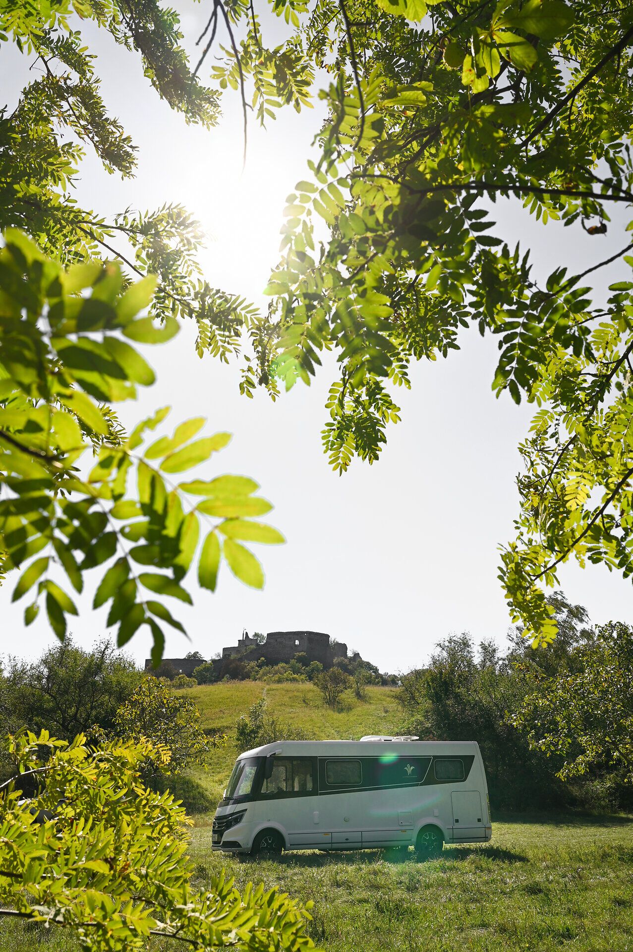 Umgeben von üppigem Grün und unter einem strahlend blauen Himmel, bietet der Campingwagen einen perfekten Rückzugsort für Naturliebhaber. Die sanften Hügel im Hintergrund und die historische Burgruine schaffen eine malerische Kulisse, die zum Entspannen und Erkunden einlädt.