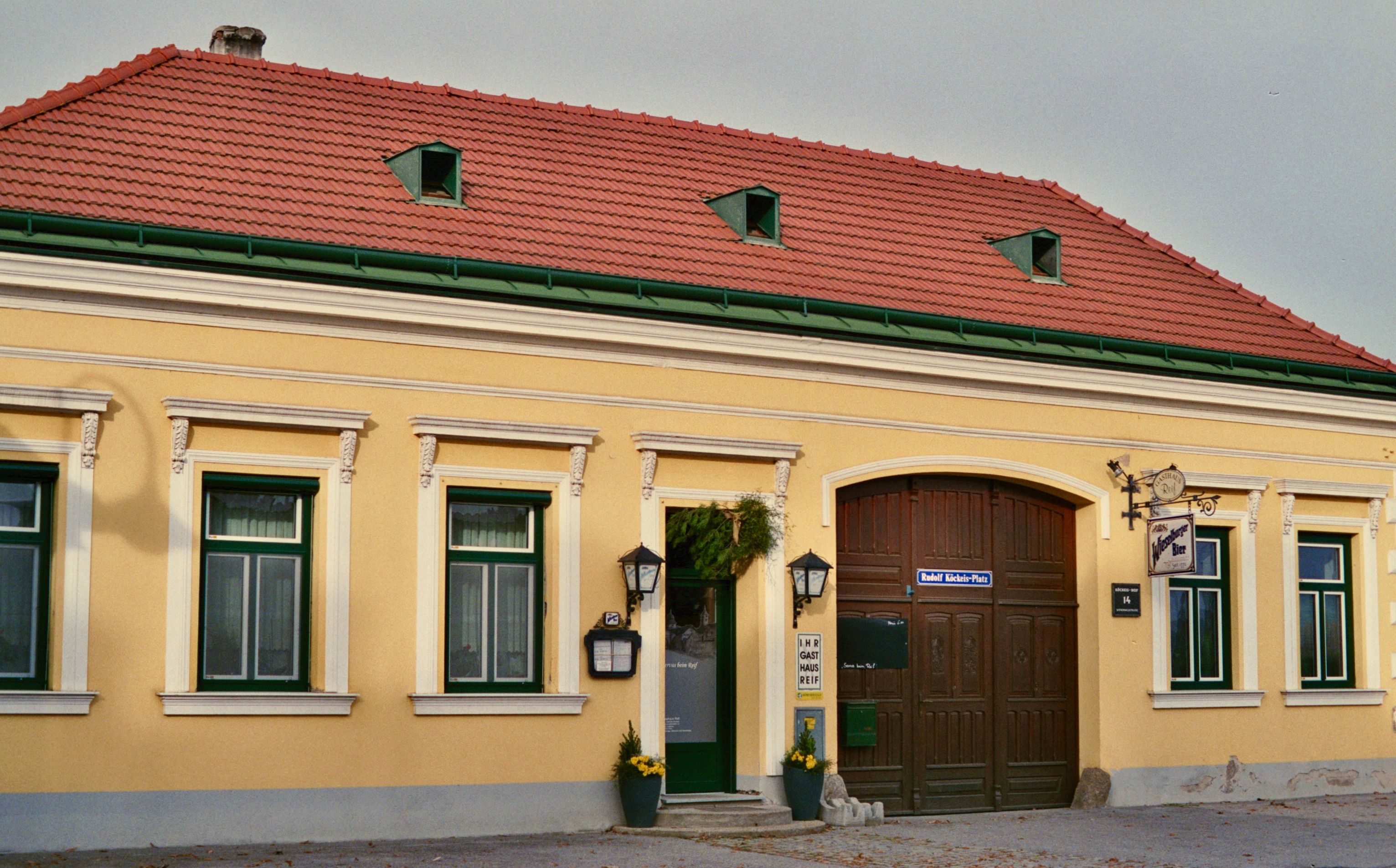 Exteriérový pohľad na žltý hostinec s červenými škridlami a zelenými okennými rámami.